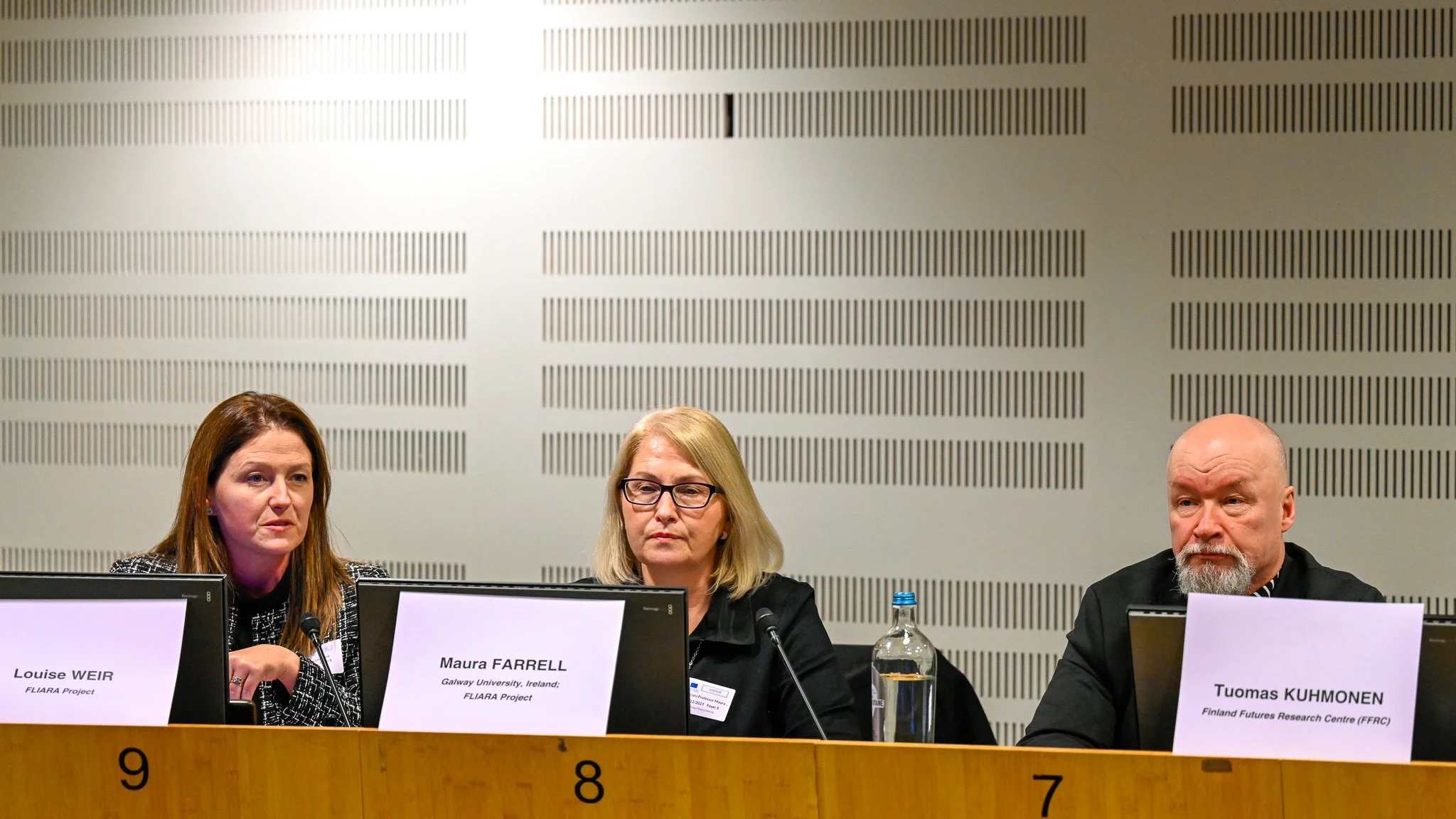 Trois personnes assises derrière des ordinateurs portables avec leurs noms visibles sur des plaques. Elles sont dans une salle de conférence.