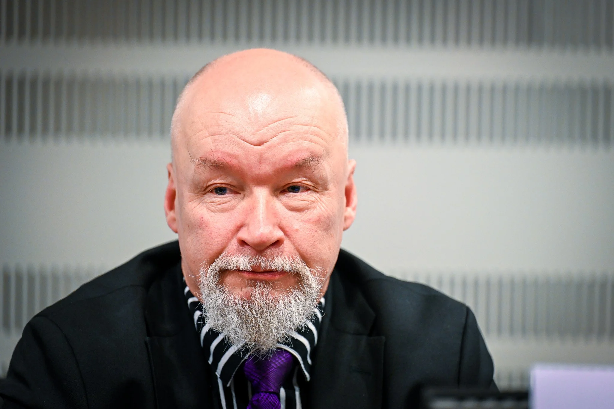 Un homme chauve portant une barbe blanche, habillé en costume noir avec une chemise à rayures et une cravate violette, regarde dans une direction indéterminée.