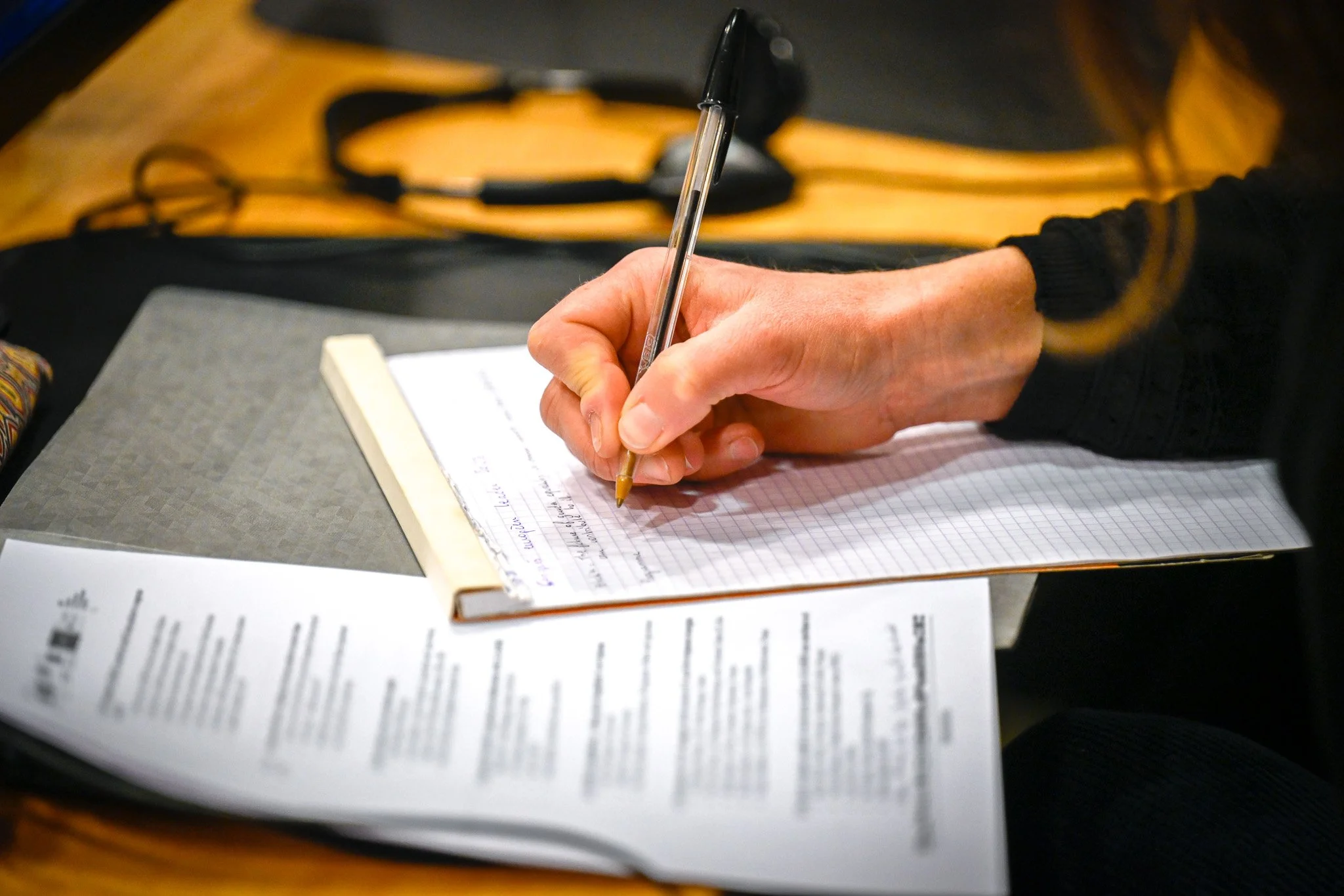 Personne écrivant avec un stylo sur un cahier quadrillé, documents à côté sur une table en bois, casque audio en arrière-plan.