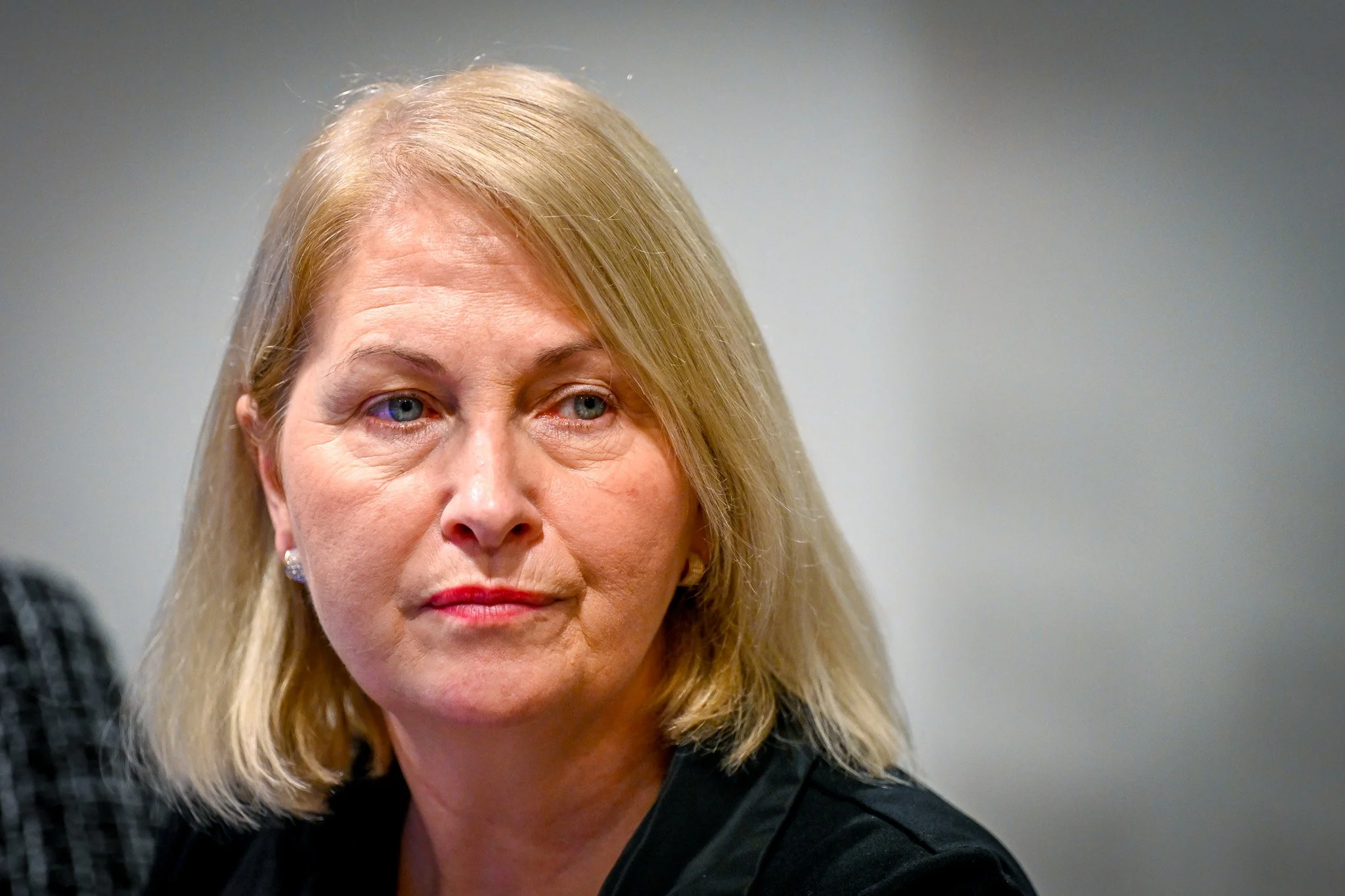 Une personne aux cheveux blonds, habillée en noir, avec une expression sérieuse.