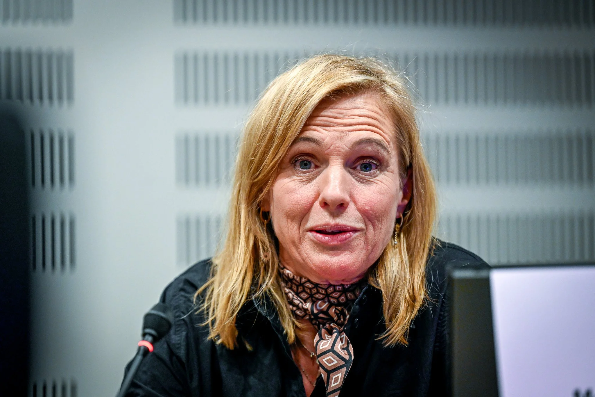 Une femme blonde portant un foulard et une chemise noire, assise devant un micro, semble parler ou être en discussion dans une salle de conférence.