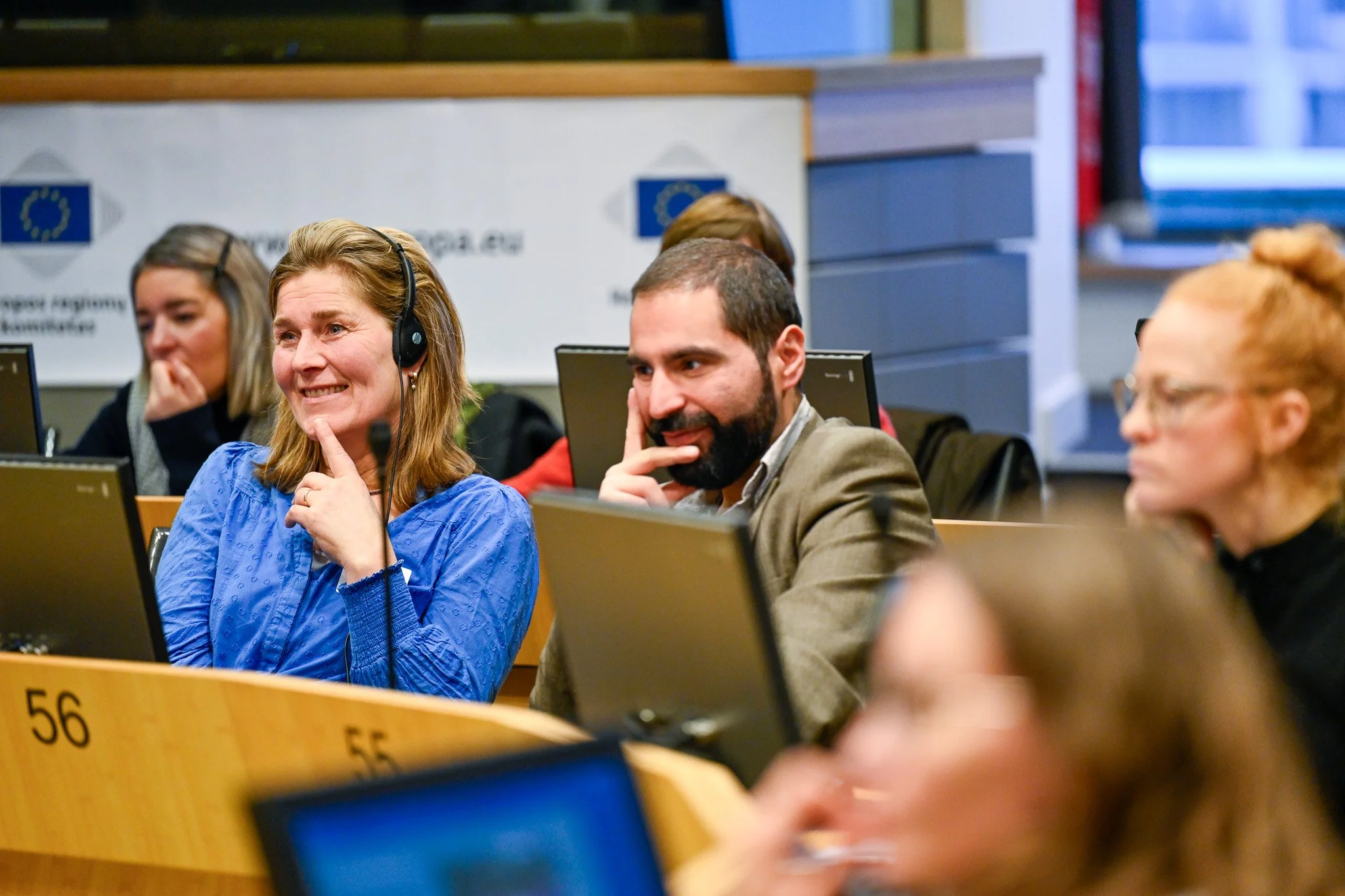Plusieurs personnes assises dans une salle de conférence, certaines utilisent des ordinateurs portables, fond avec un logo de l'Union européenne.