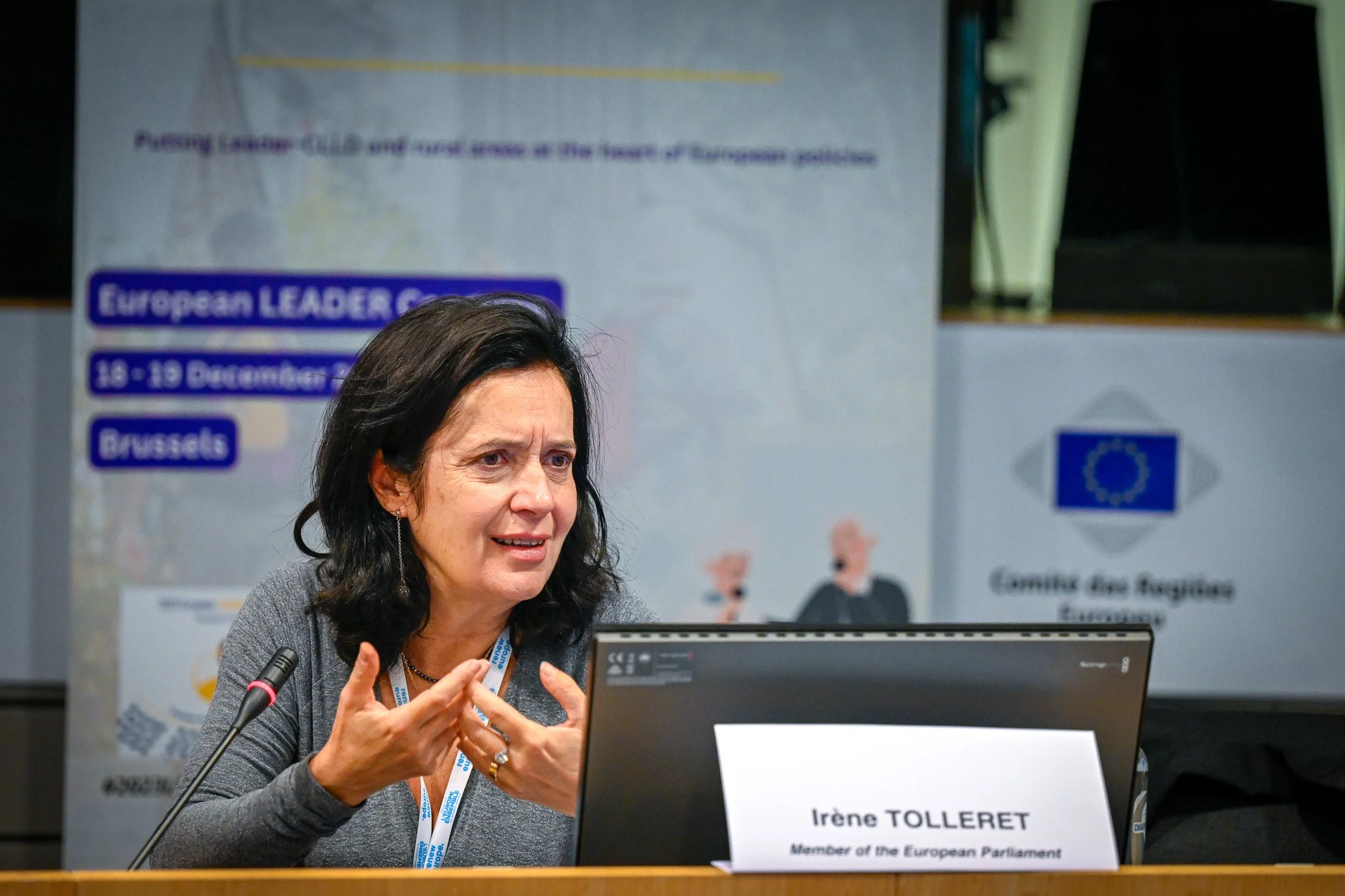 Femme s'exprimant lors d'une conférence à Bruxelles, avec un panneau indiquant "Membre du Parlement Européen" devant elle, fond avec des logos européens.
