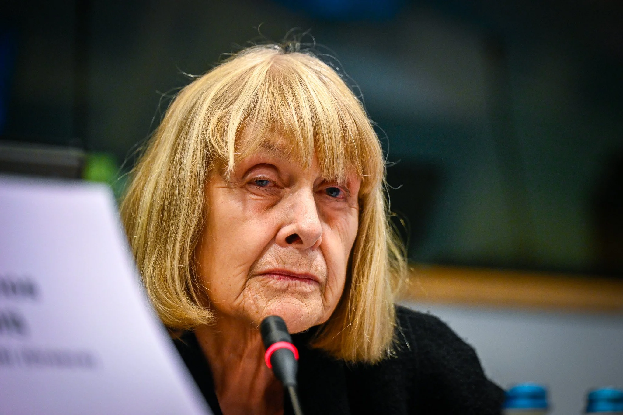 Une femme âgée avec des cheveux blonds, assise derrière un micro lors d'un événement ou une conférence.