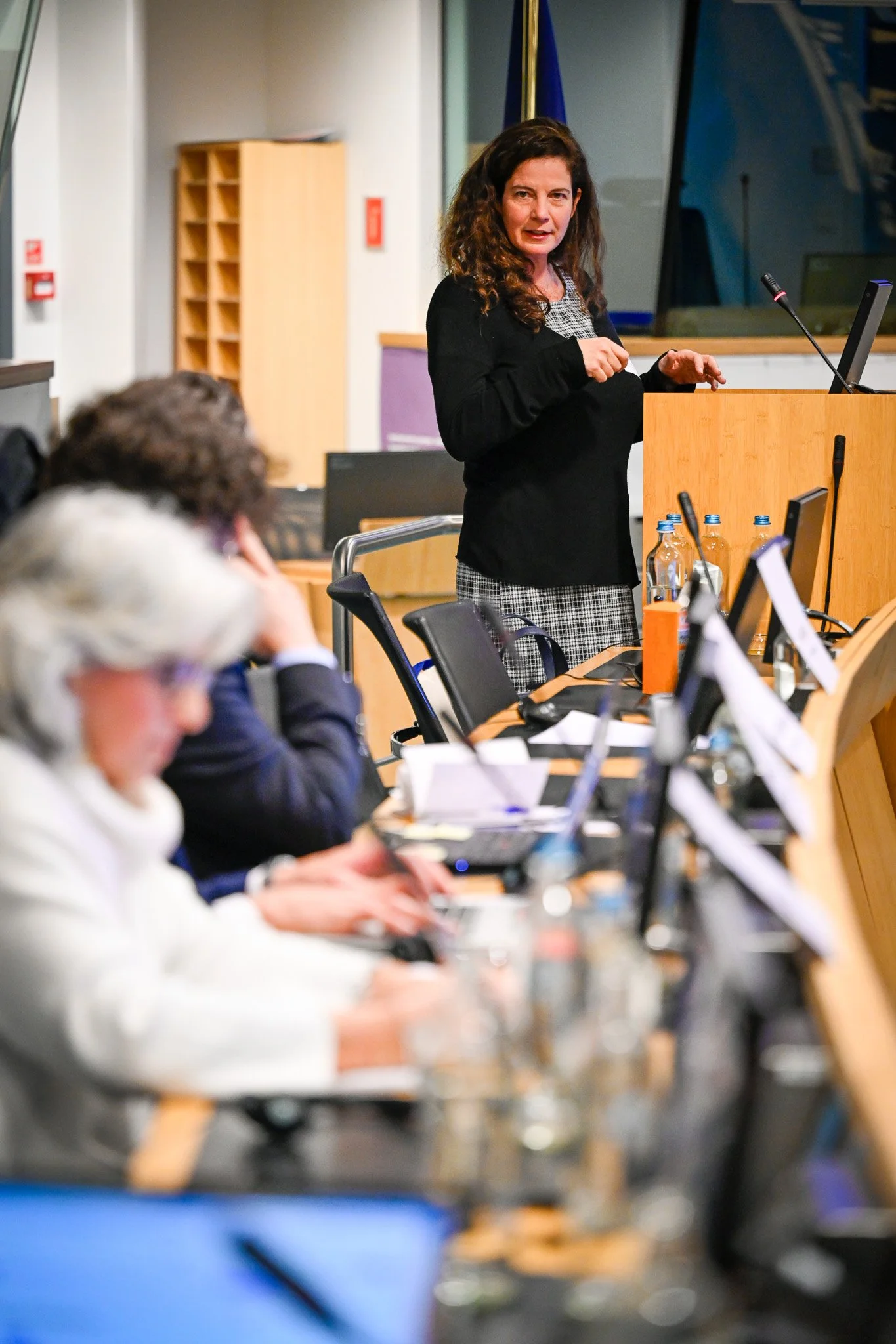 Une femme en train de faire une présentation devant un pupitre, avec des personnes assises à une table utilisant des ordinateurs portables.