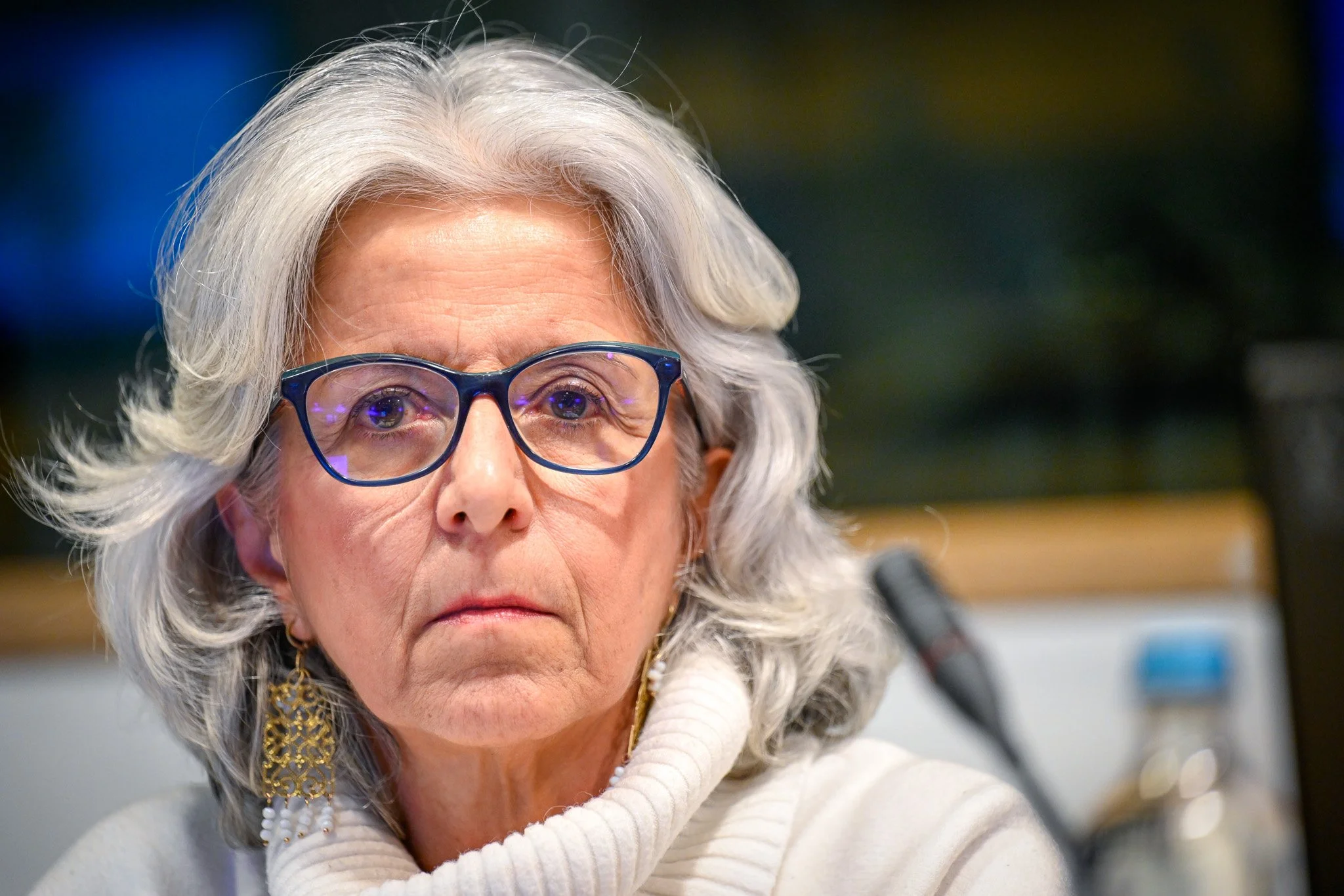 Femme aux cheveux gris, portant des lunettes, un pull blanc, et des boucles d'oreilles dorées.