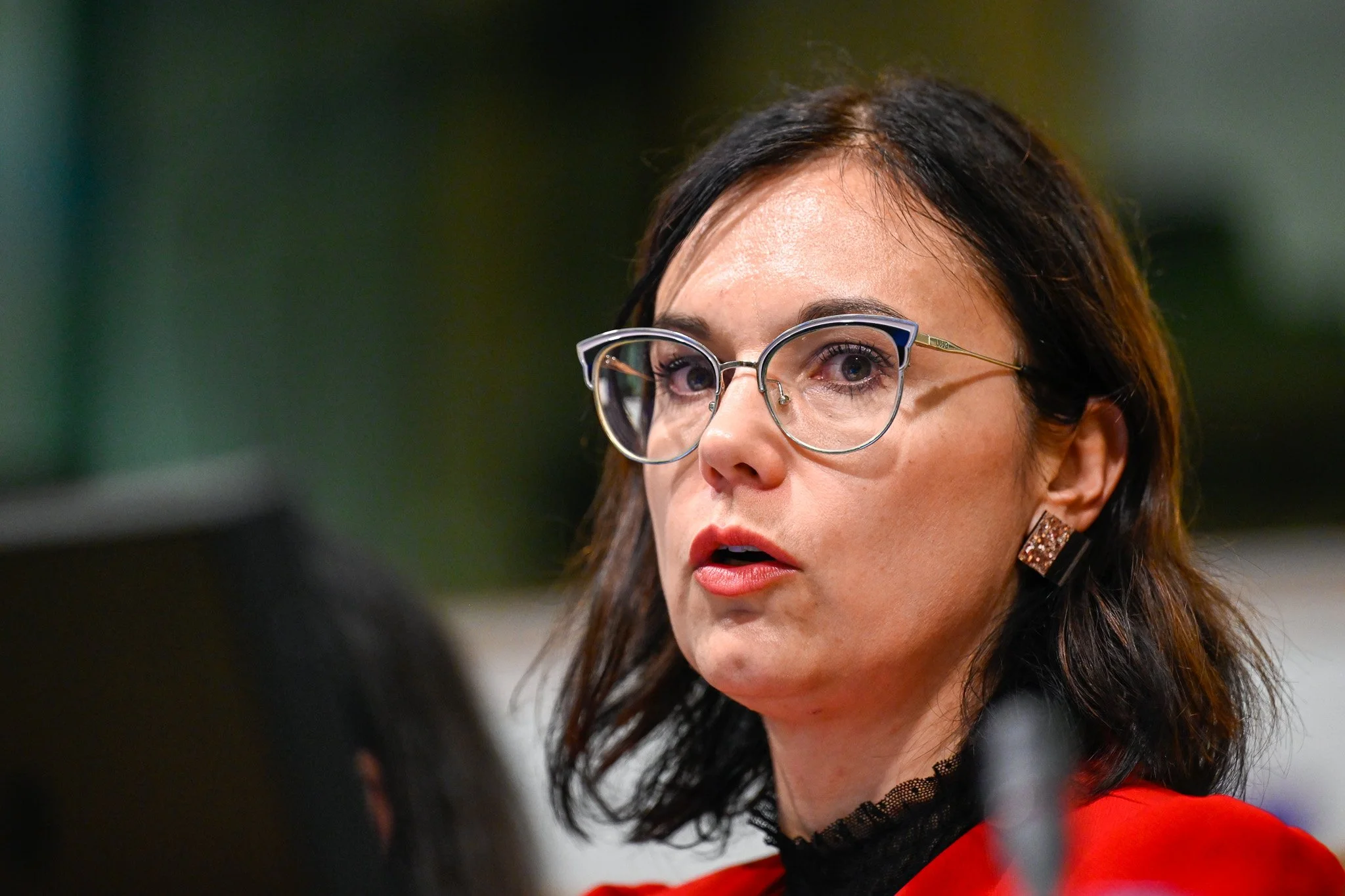Femme avec lunettes et cheveux foncés, portant un rouge à lèvres rouge, prise lors d'un discours ou d'une présentation.