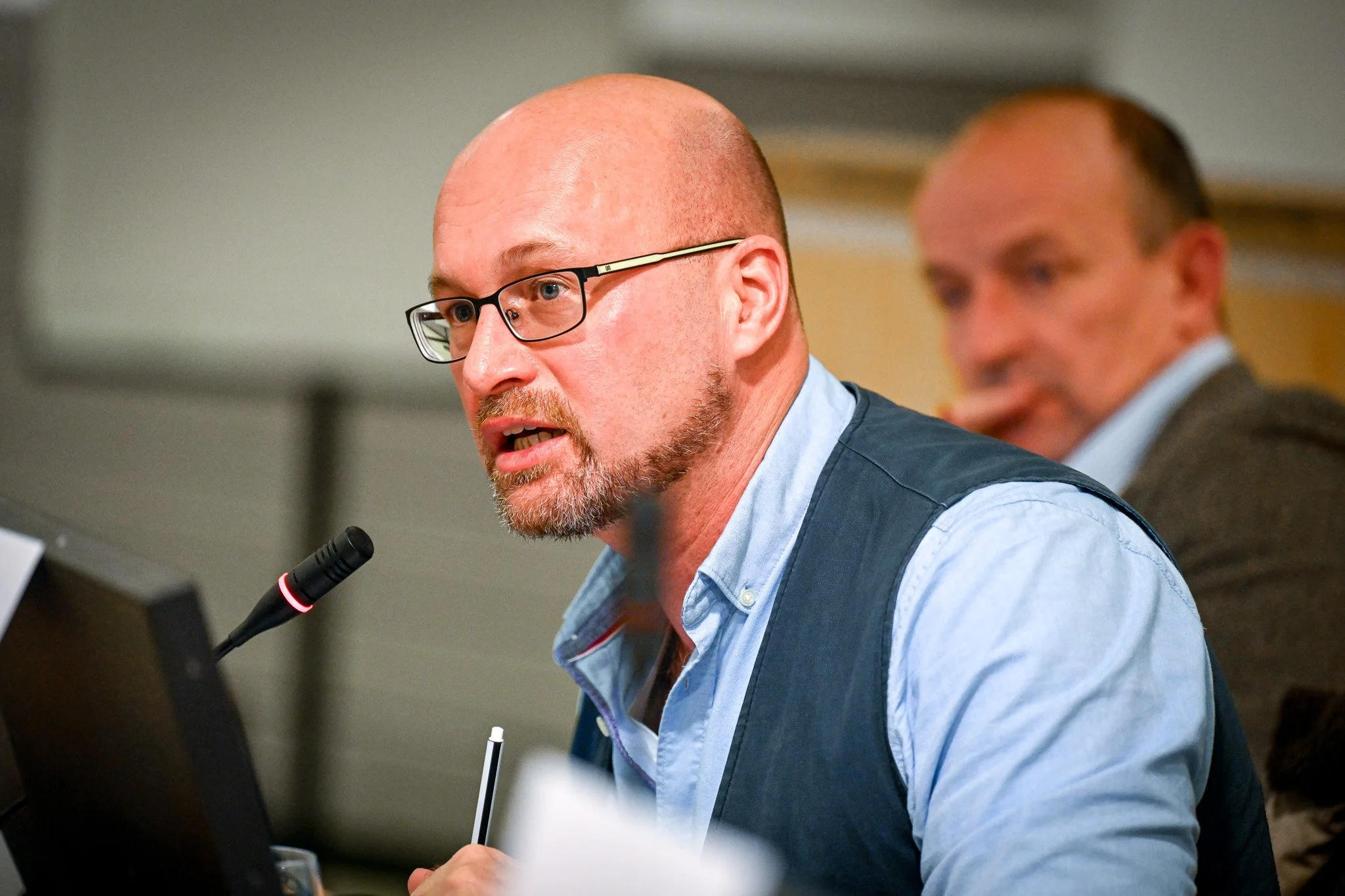 Un homme chauve avec des lunettes parle dans un microphone lors d'une réunion ou d'une conférence. Une autre personne floue est visible en arrière-plan.