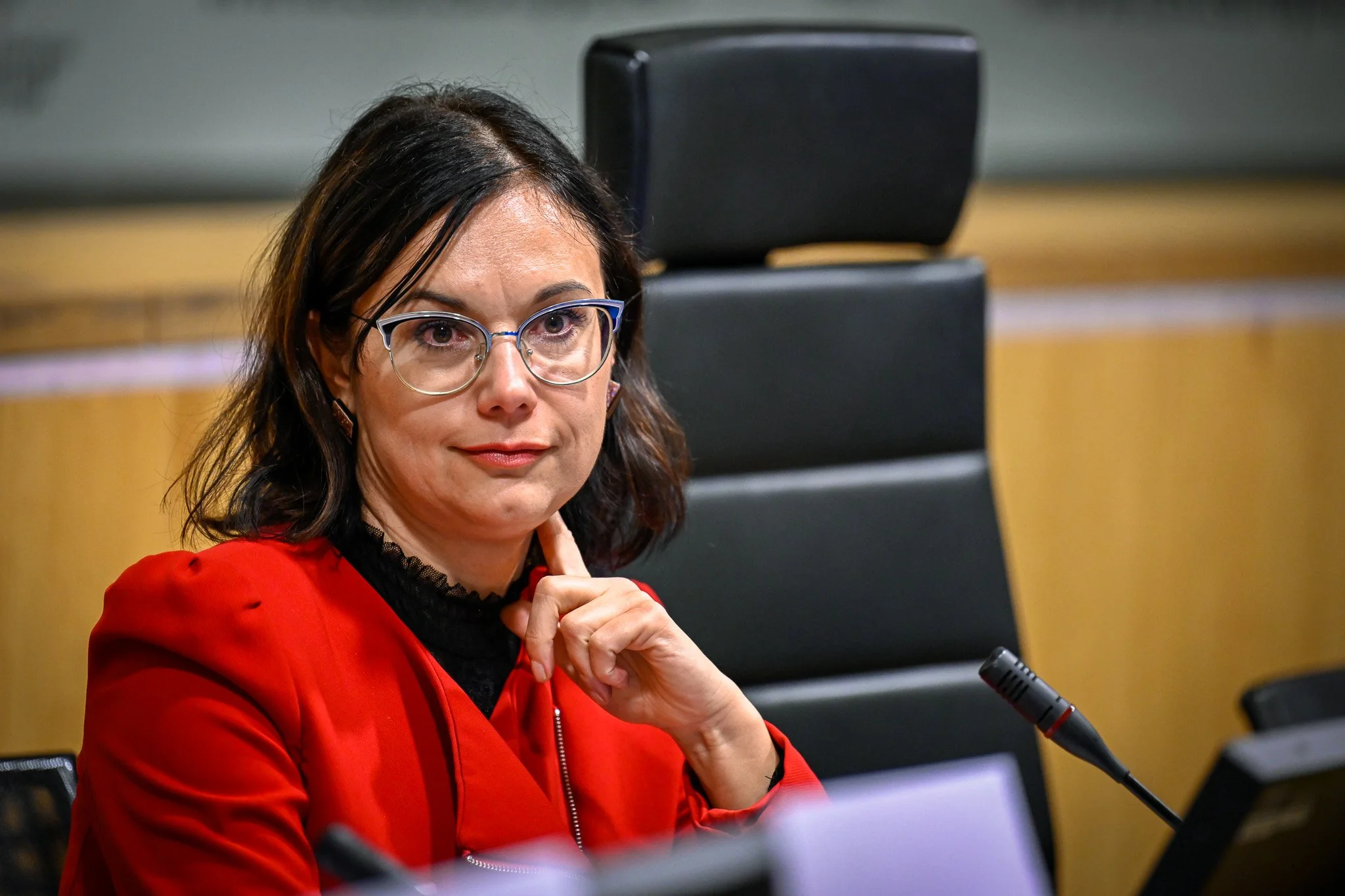 Une femme portant des lunettes et un vêtement rouge est assise devant un micro, dans une salle de conférence.