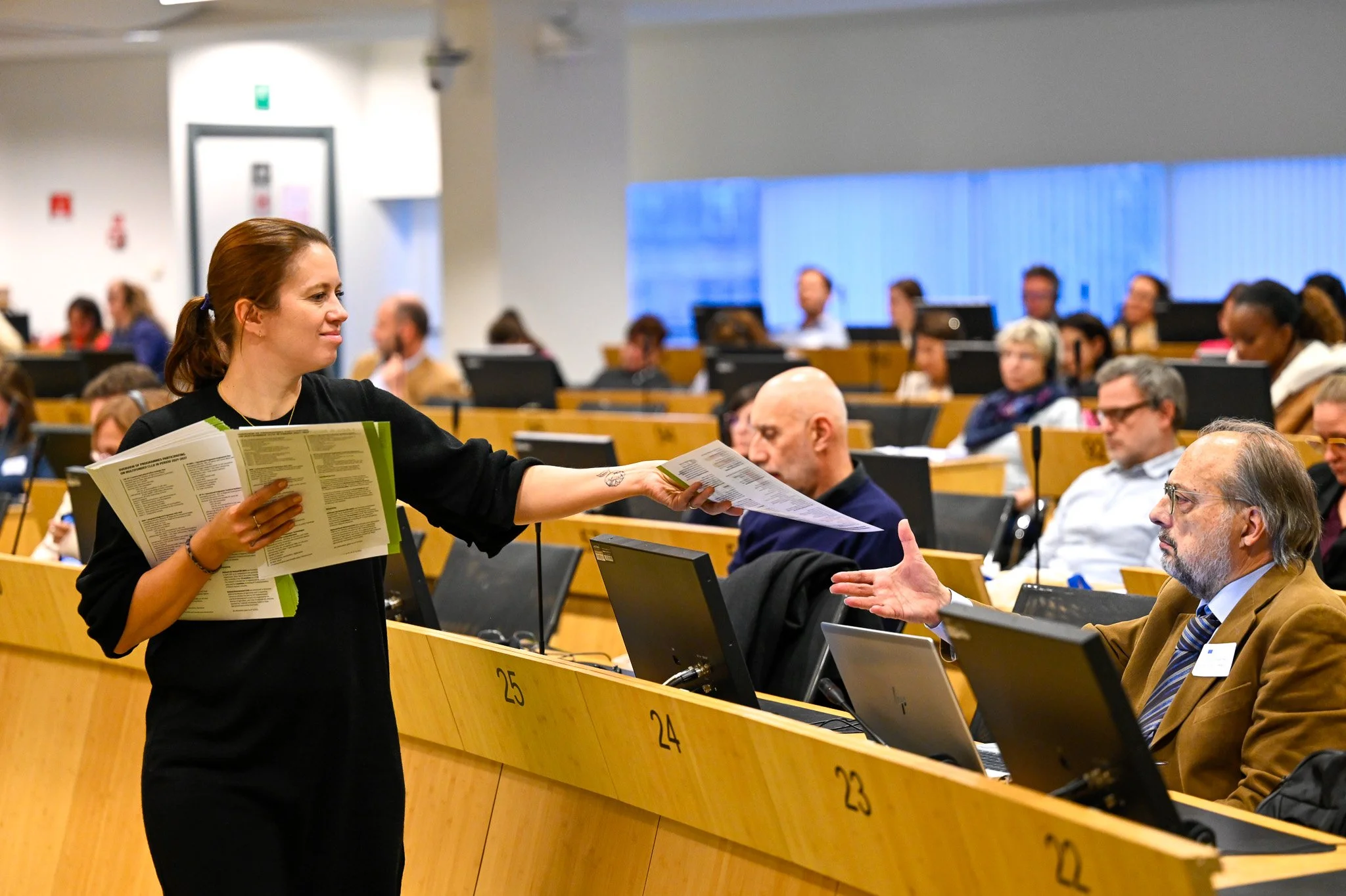 Une femme distribue des documents à un homme dans une salle de conférence avec plusieurs participants assis devant des ordinateurs portables.