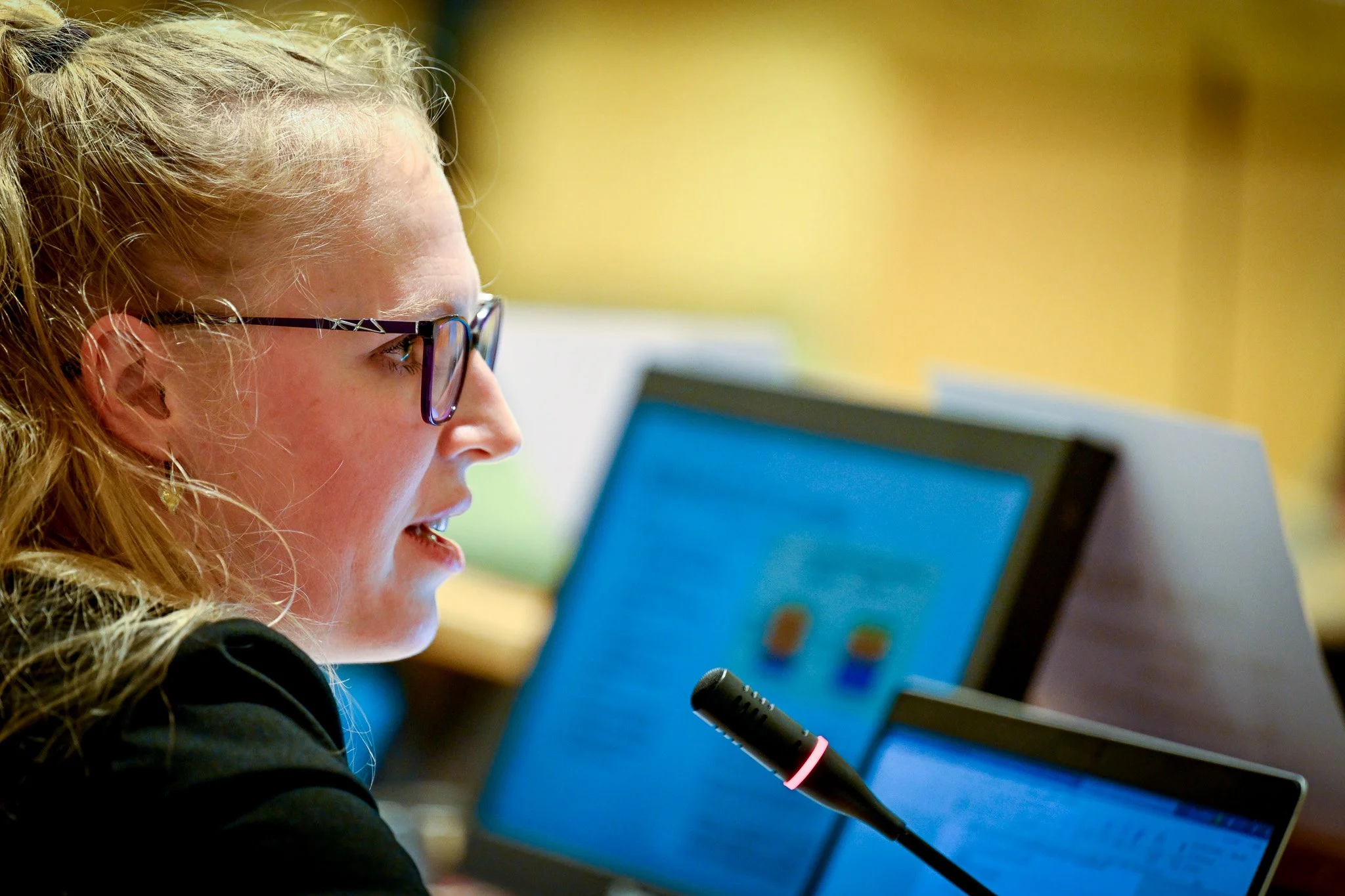 Femme portant des lunettes parlant dans un microphone avec des écrans et documents en arrière-plan.
