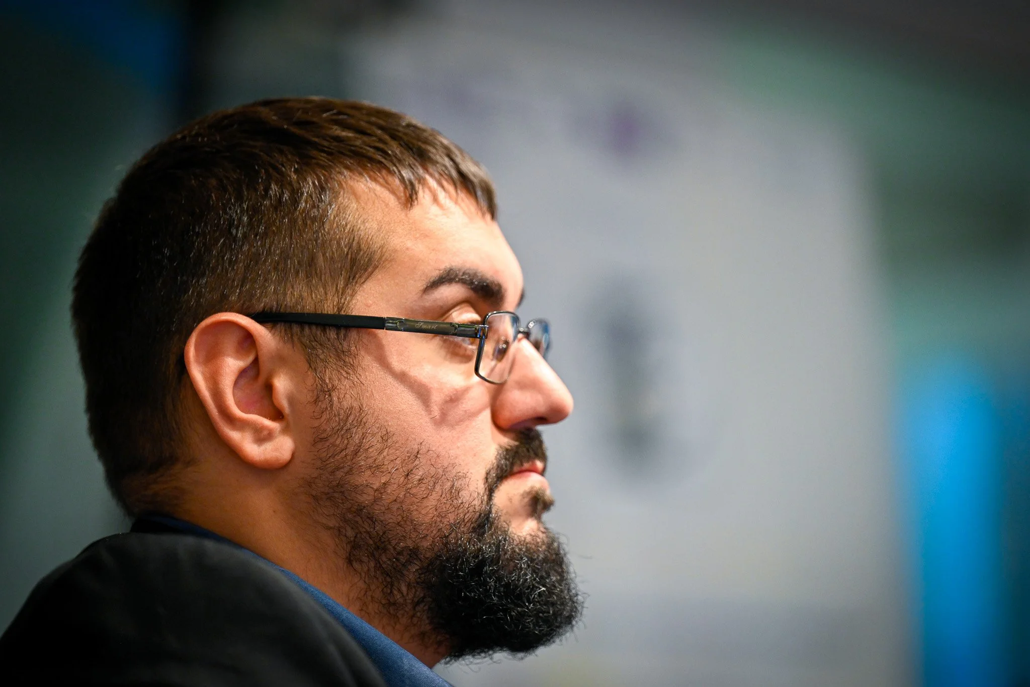 Profil d'un homme barbu portant des lunettes, regardant devant lui, fond flou.