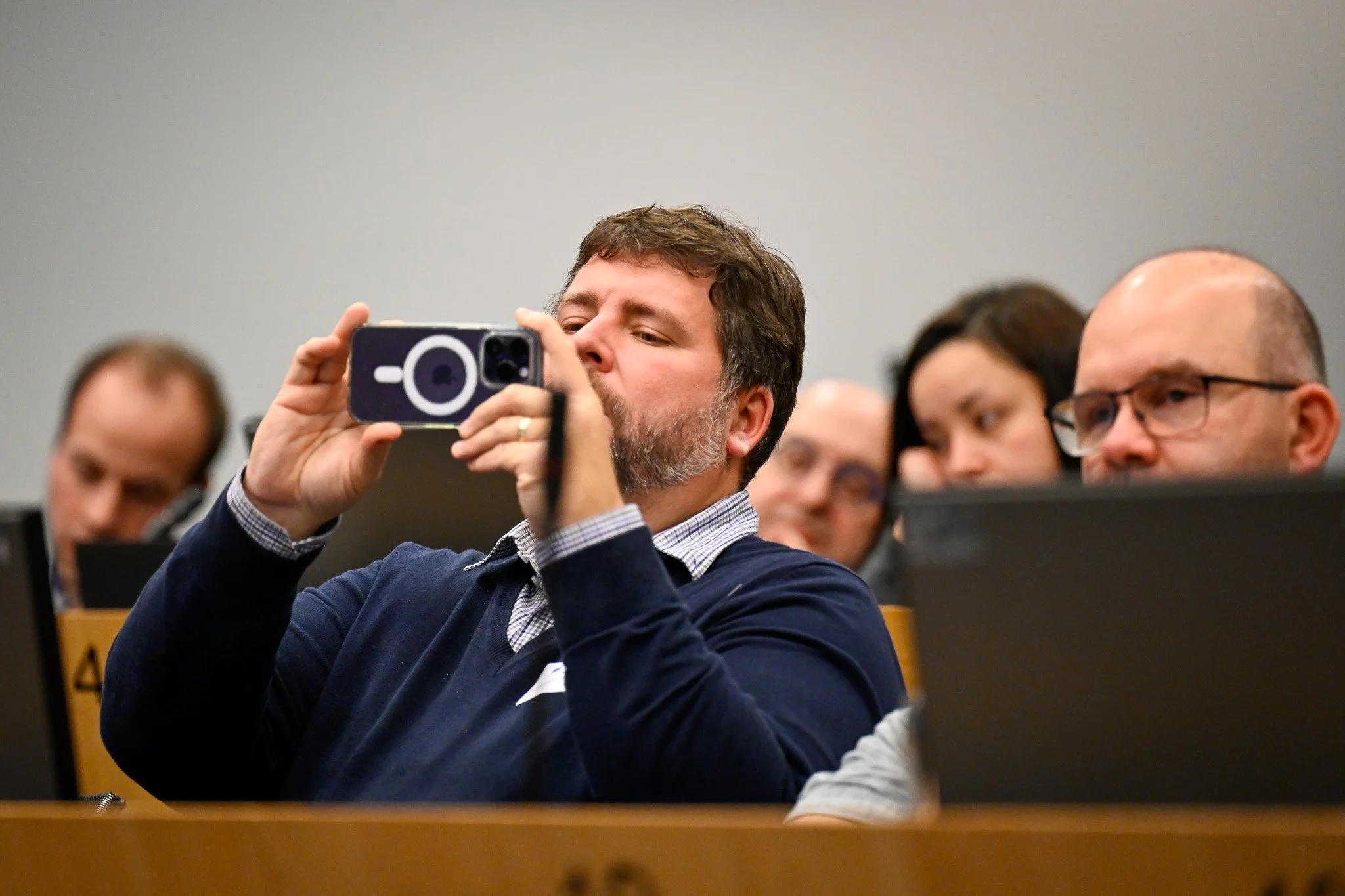 Un homme prend une photo avec un smartphone dans une salle de réunion, entouré d'autres personnes assises.