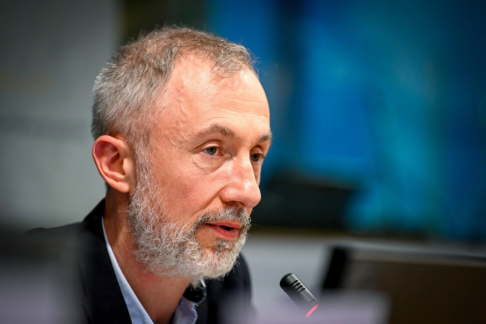 Homme âgé avec barbe parle près d'un microphone.
