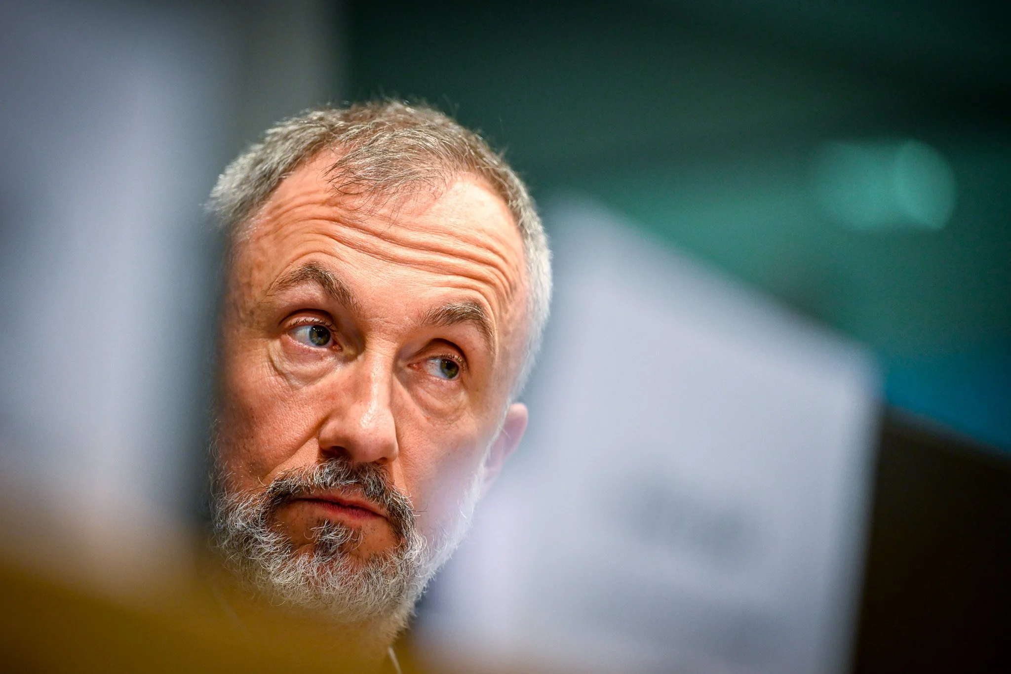 Homme avec barbe grise, yeux marron, fond flou, intérieur.
