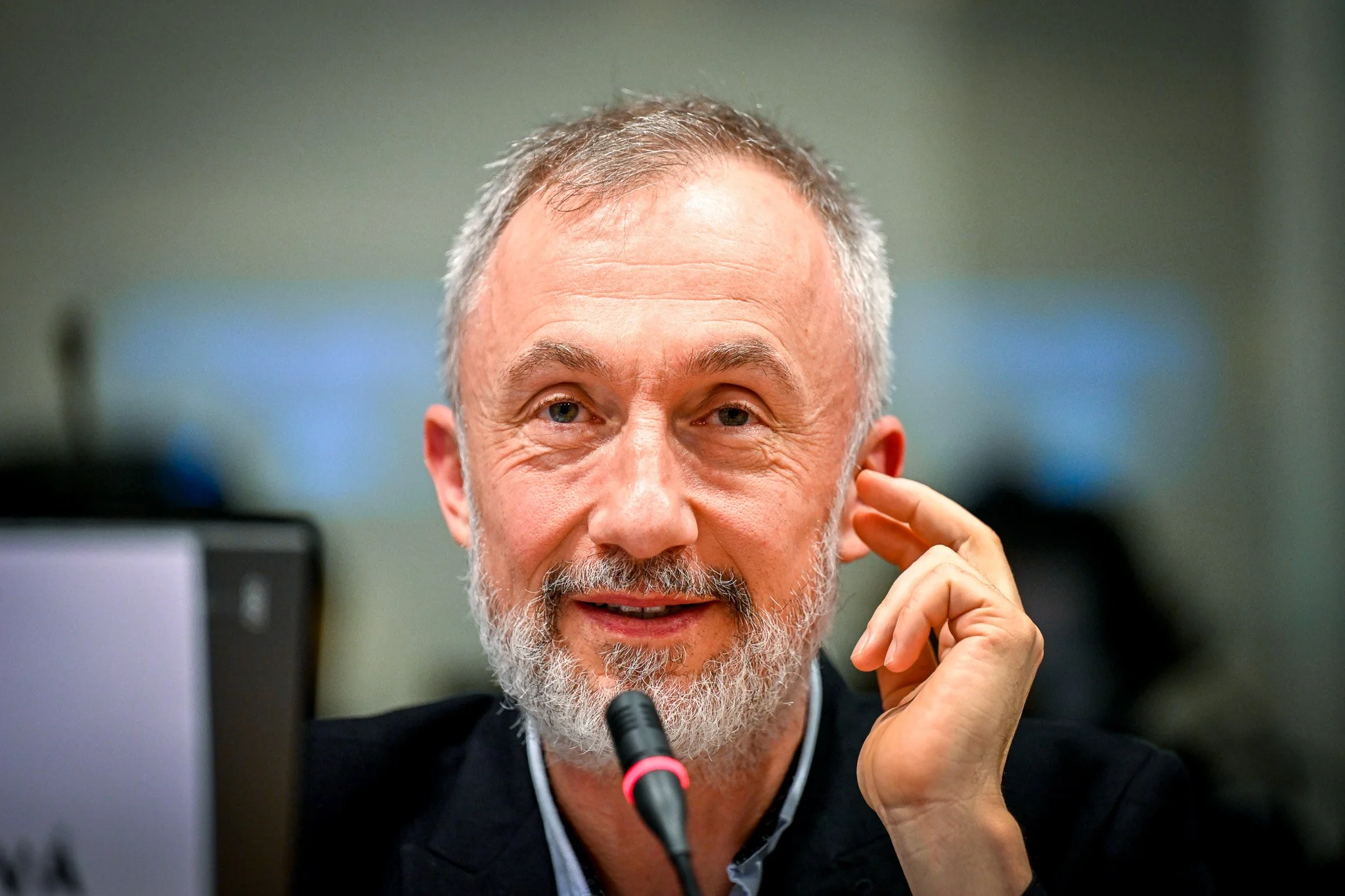 Homme âgé avec barbe grise parlant lors d'une réunion, assis devant un micro.