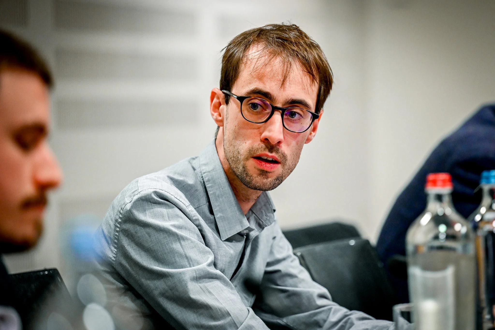 Un homme avec des lunettes assis à une table, portant une chemise grise, avec des bouteilles d'eau en premier plan.