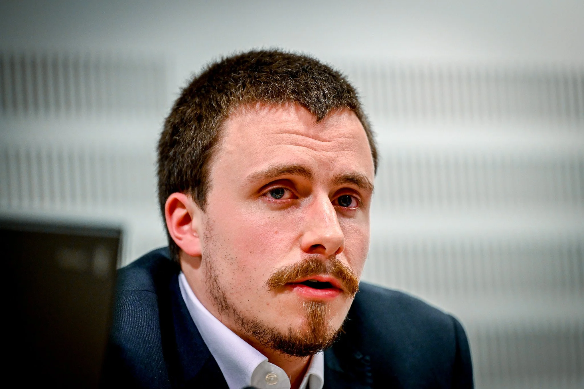 Un homme en costume avec une barbe et une moustache, regardant vers l'avant, sur un fond neutre.