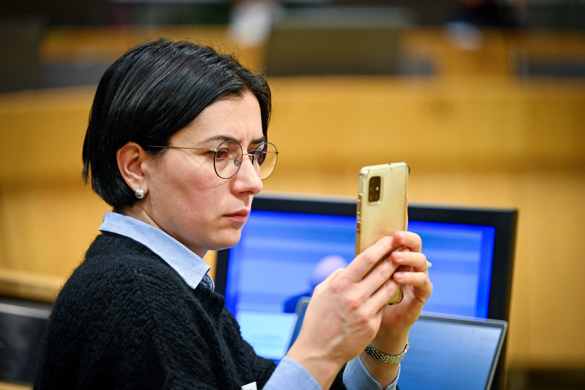Femme avec des lunettes utilisant un smartphone, un écran d'ordinateur en arrière-plan.