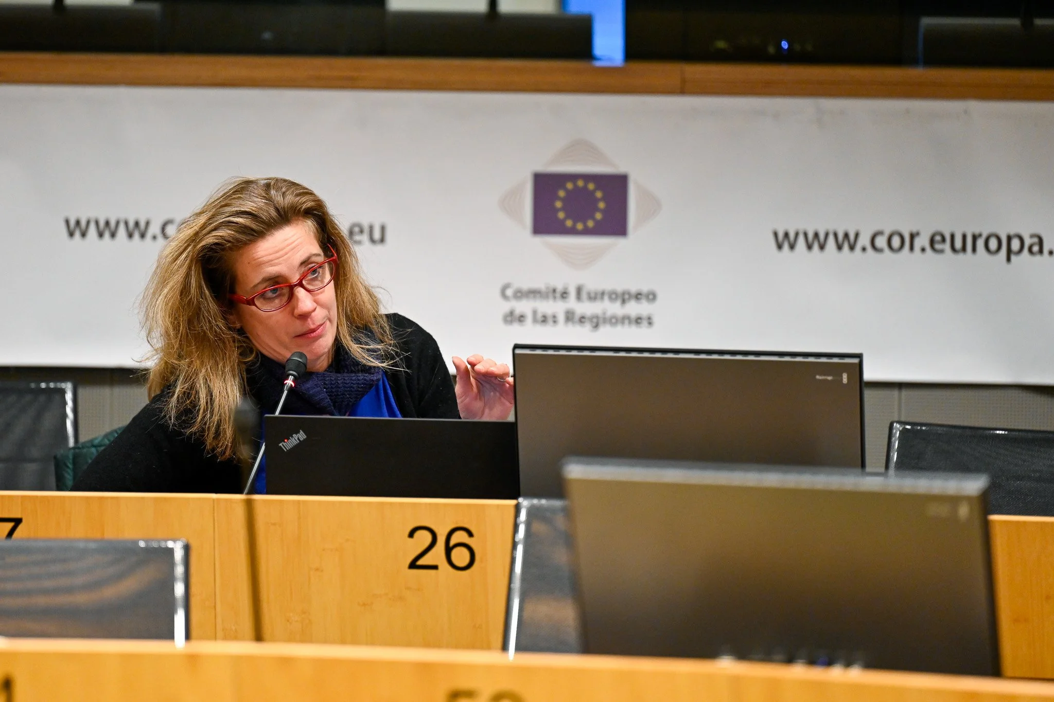 Une femme avec des lunettes parle dans un micro lors d'une réunion au Comité Européen des Régions. Derrière elle, un panneau montre le logo et le site web du comité.