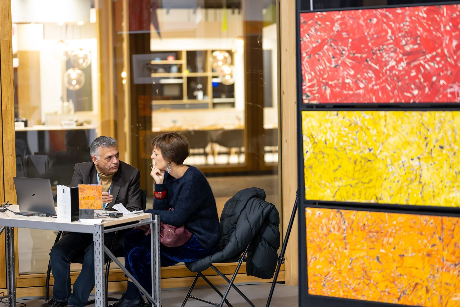 Vincent Parissis and Élodie Treillard are chatting at a table in the ART NFT Gallery, surrounded by red and yellow artworks.