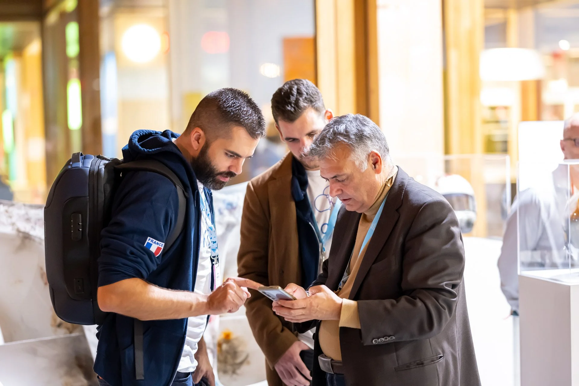 Three men looking at a mobile phone in a brightly lit indoor setting.