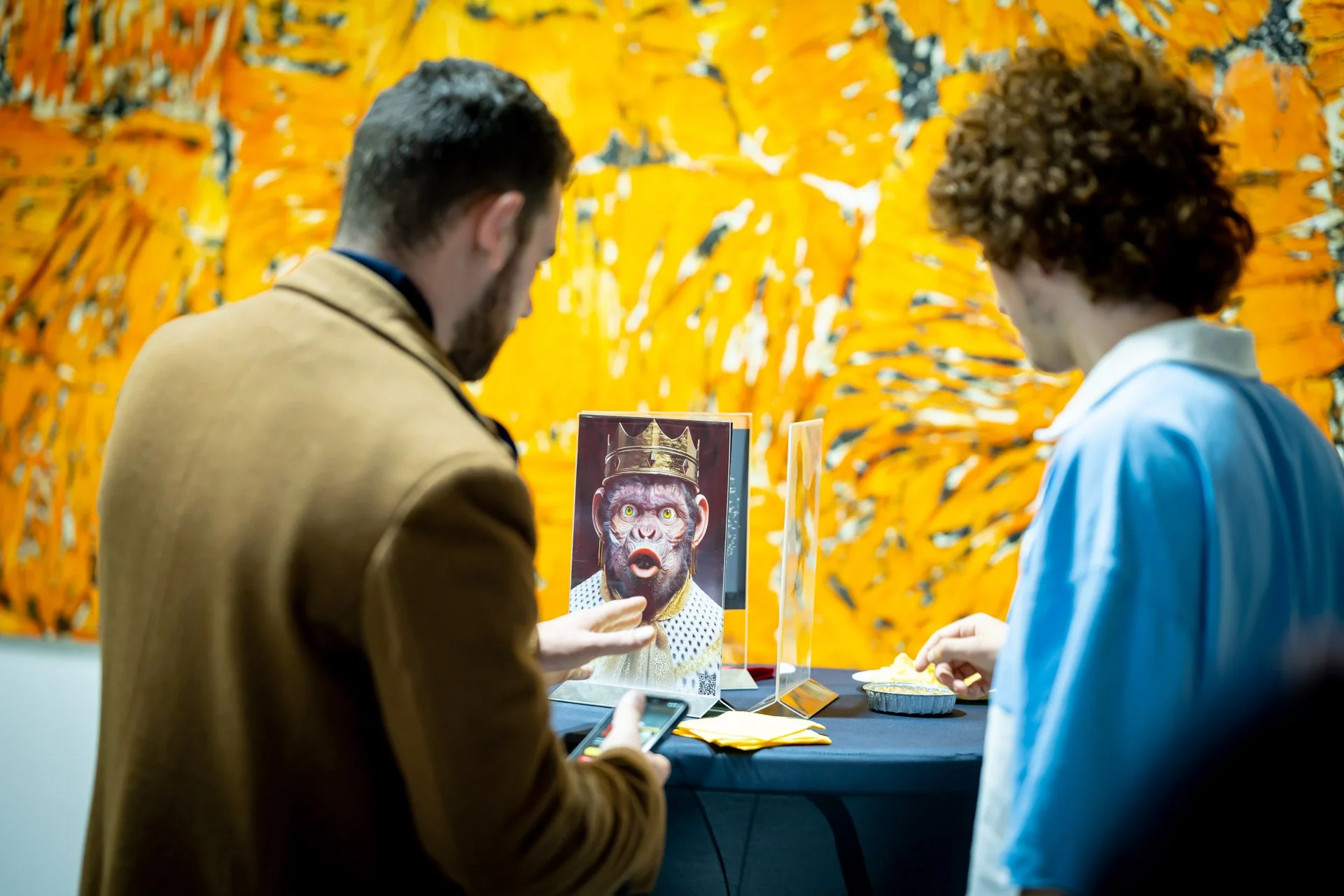 Two visitors are looking at a screen at a table in the ART NFT Gallery, in front of a piece featuring shades of yellow and orange.