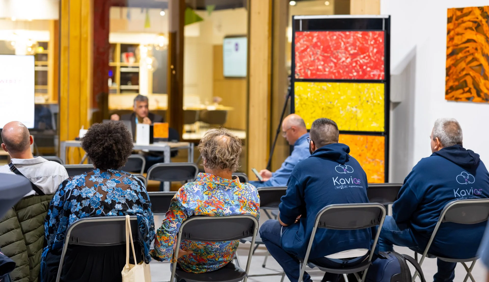 People attending a presentation in a room with colourful abstract artworks on the walls and someone speaking at the front.