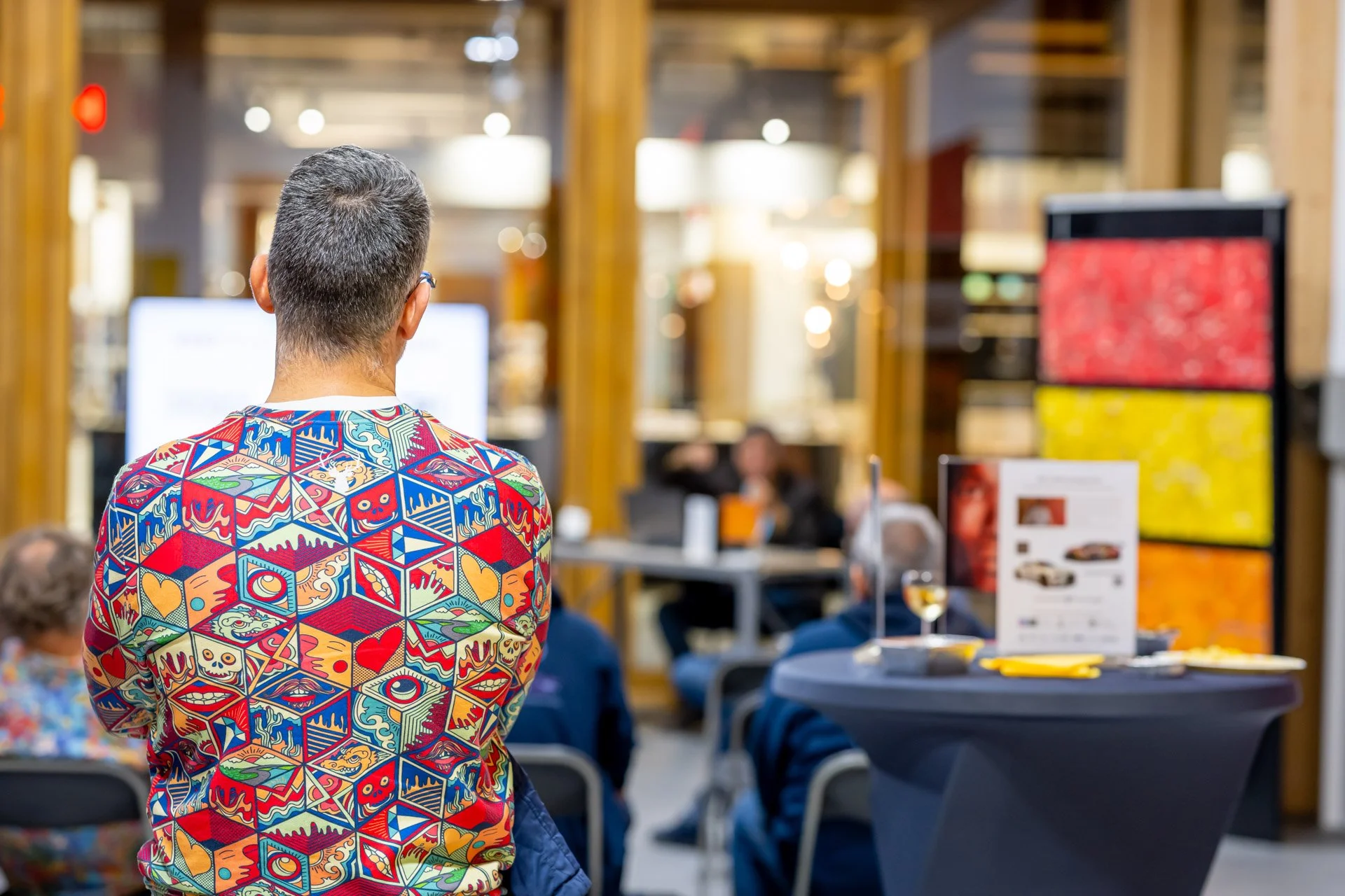 A person wearing a colourful jumper is standing with their back to the camera, taking part in an event or meeting in a modern indoor space with chairs and displays in the background.
