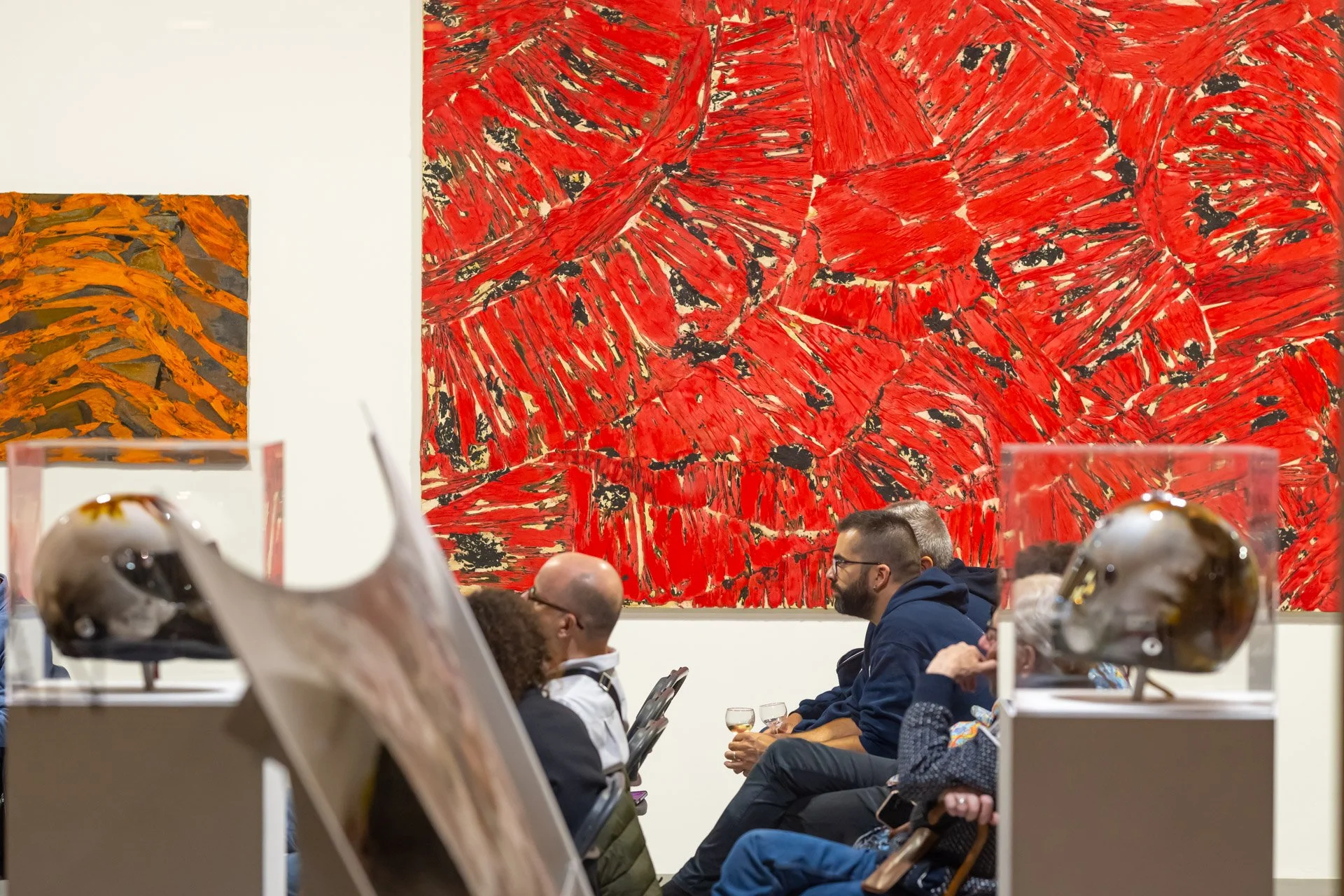 People sitting in front of red and orange abstract paintings in an art gallery, with glass sculptures on plinths in the foreground.