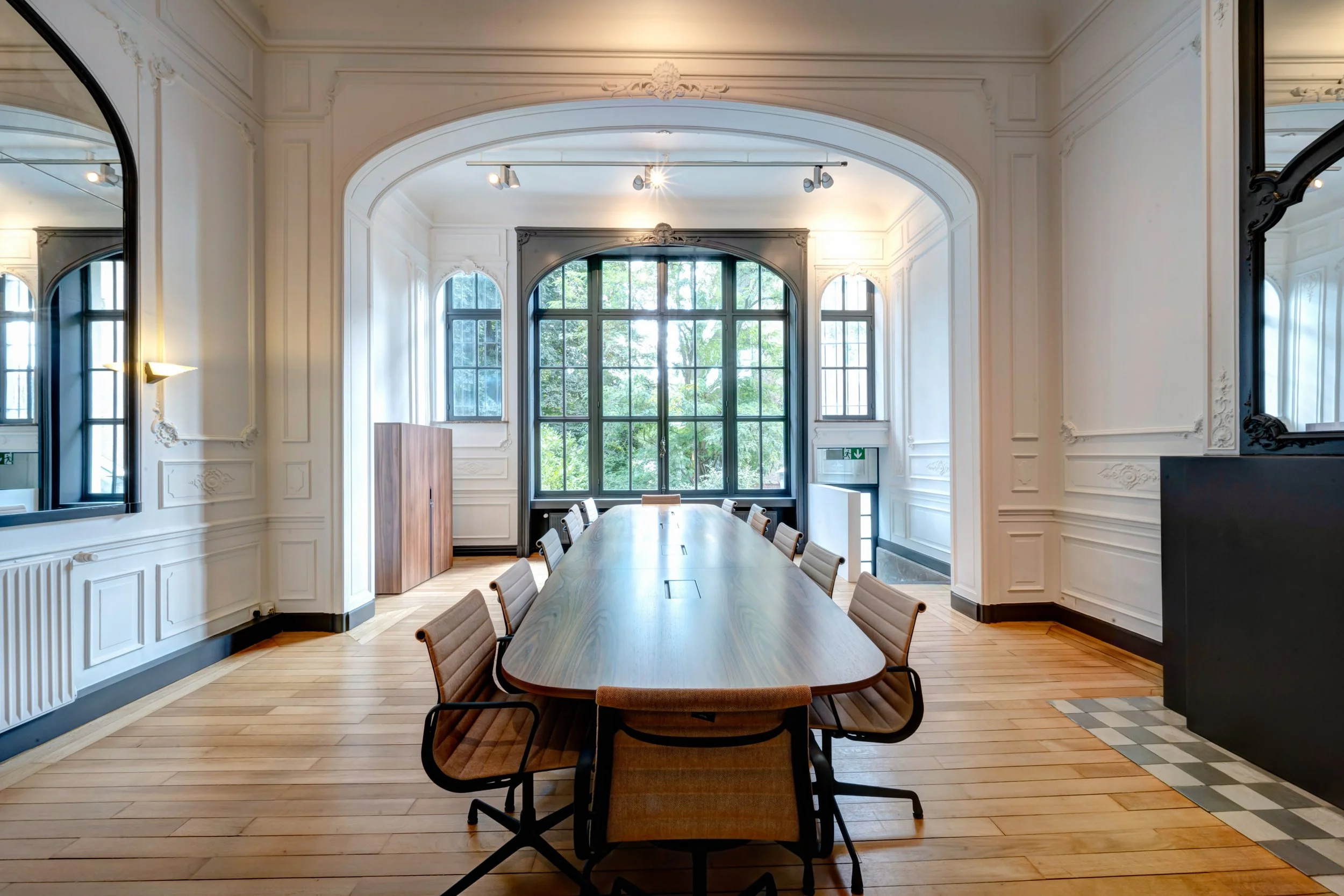 Salle de réunion avec une grande table en bois, chaises en tissu beige, grandes fenêtres laissant entrer la lumière naturelle, murs blanc avec moulures, sol en bois, et miroir noir