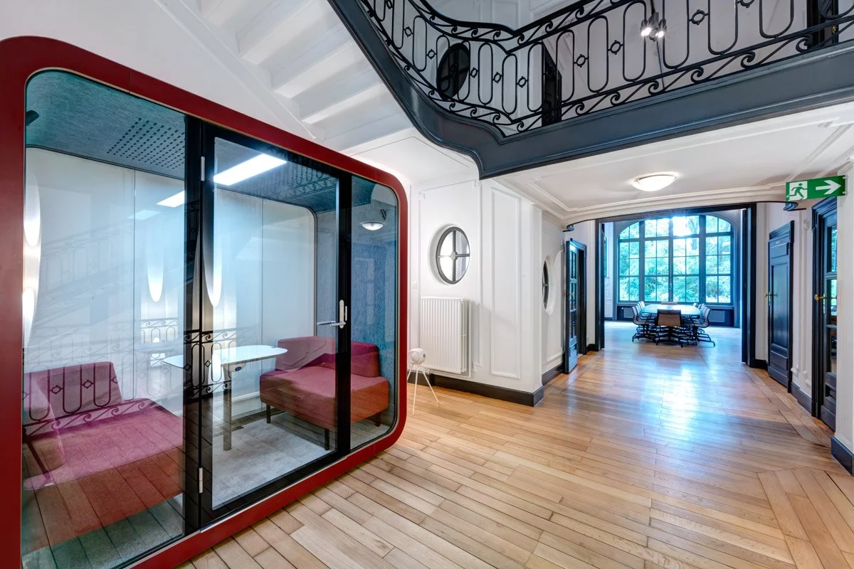 Un espace intérieur avec parquet, une cabine vitrée à cadre rouge et un escalier à balustrade noire.