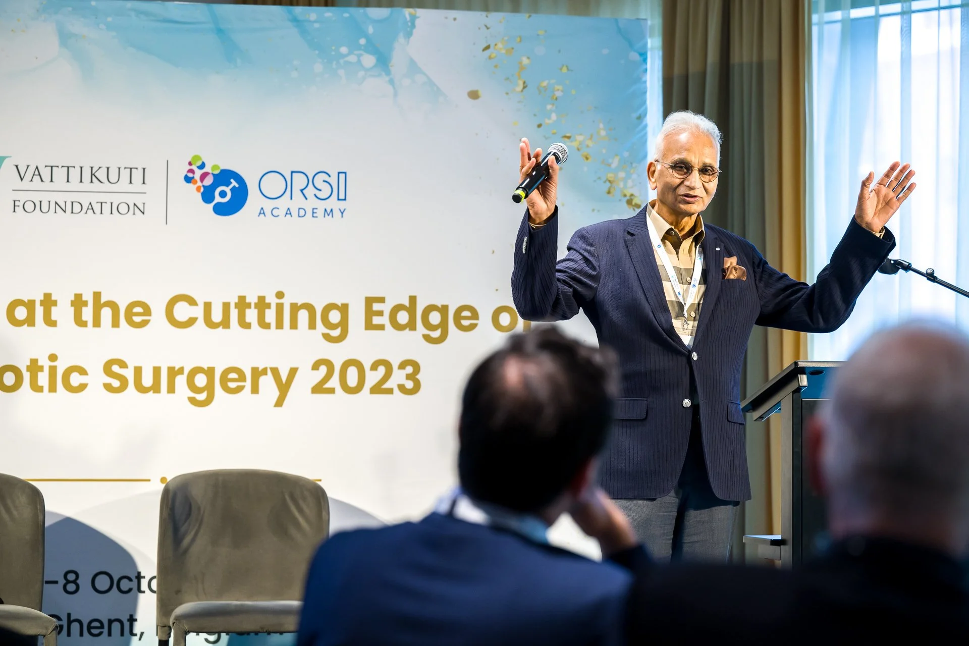 Un homme en costume tient un micro et parle lors d'une conférence sur la chirurgie robotique en 2023. Il se tient devant une affiche avec les logos de la Fondation Vattikuti et de l'Académie ORSI.