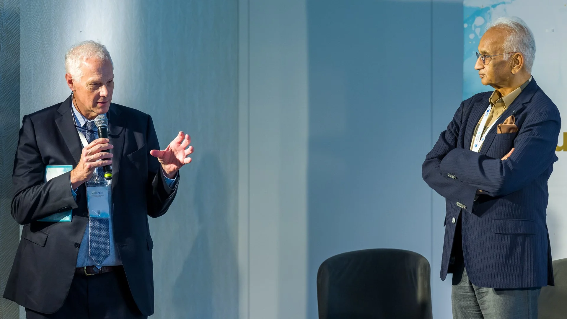 Deux hommes habillés en costumes discutant sur une scène lors d'une conférence, l'un d'eux tenant un microphone.