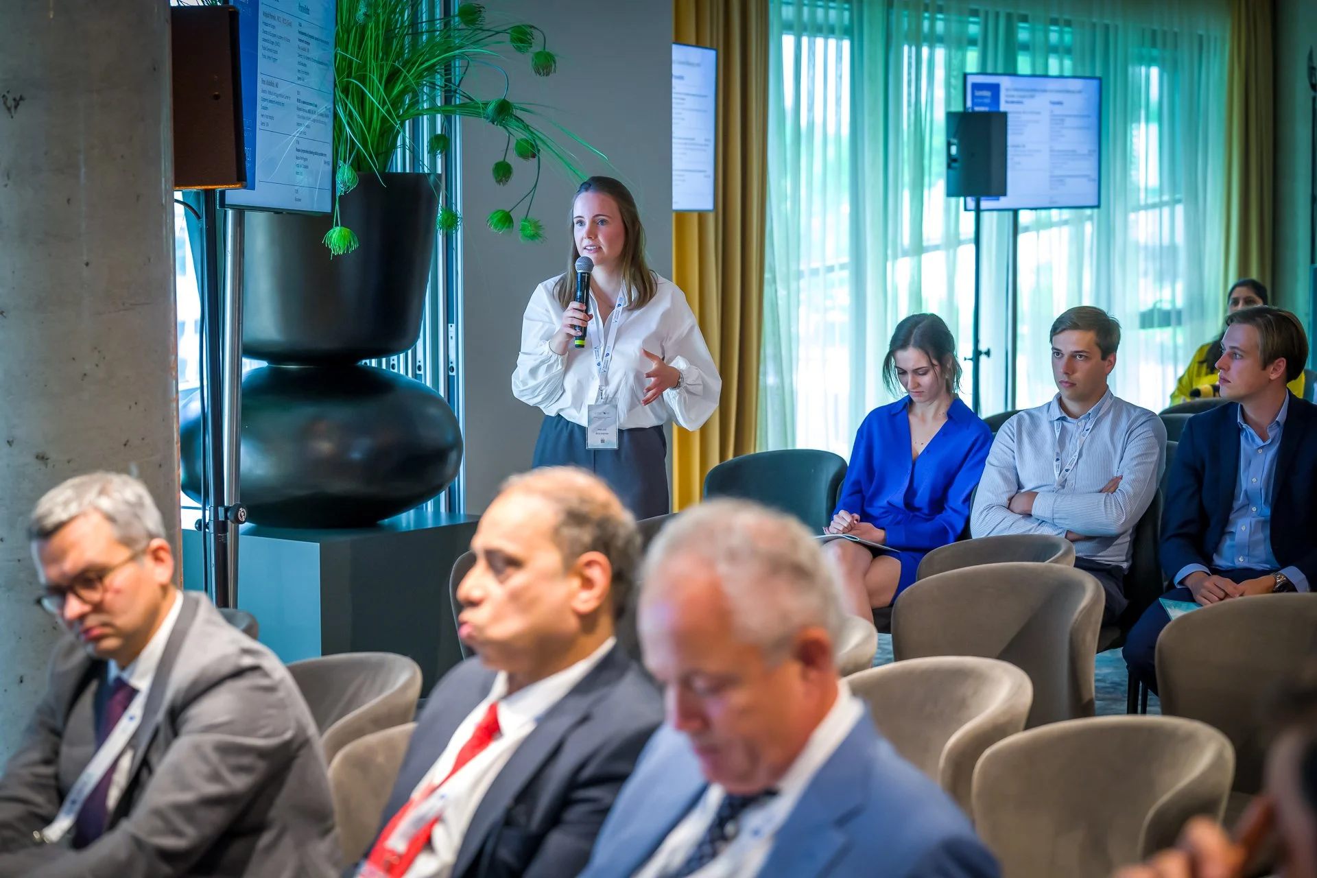 Une personne parle au micro lors d'une conférence, tandis que plusieurs participants sont assis et écoutent. La salle est équipée d'écrans et de décorations modernes.