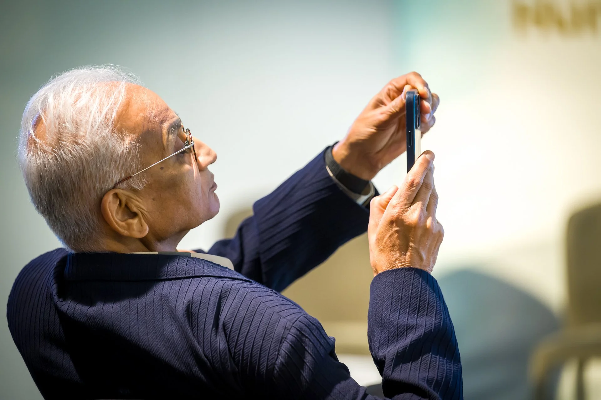Un homme aux cheveux gris, portant des lunettes et un costume, utilise un smartphone pour prendre une photo.