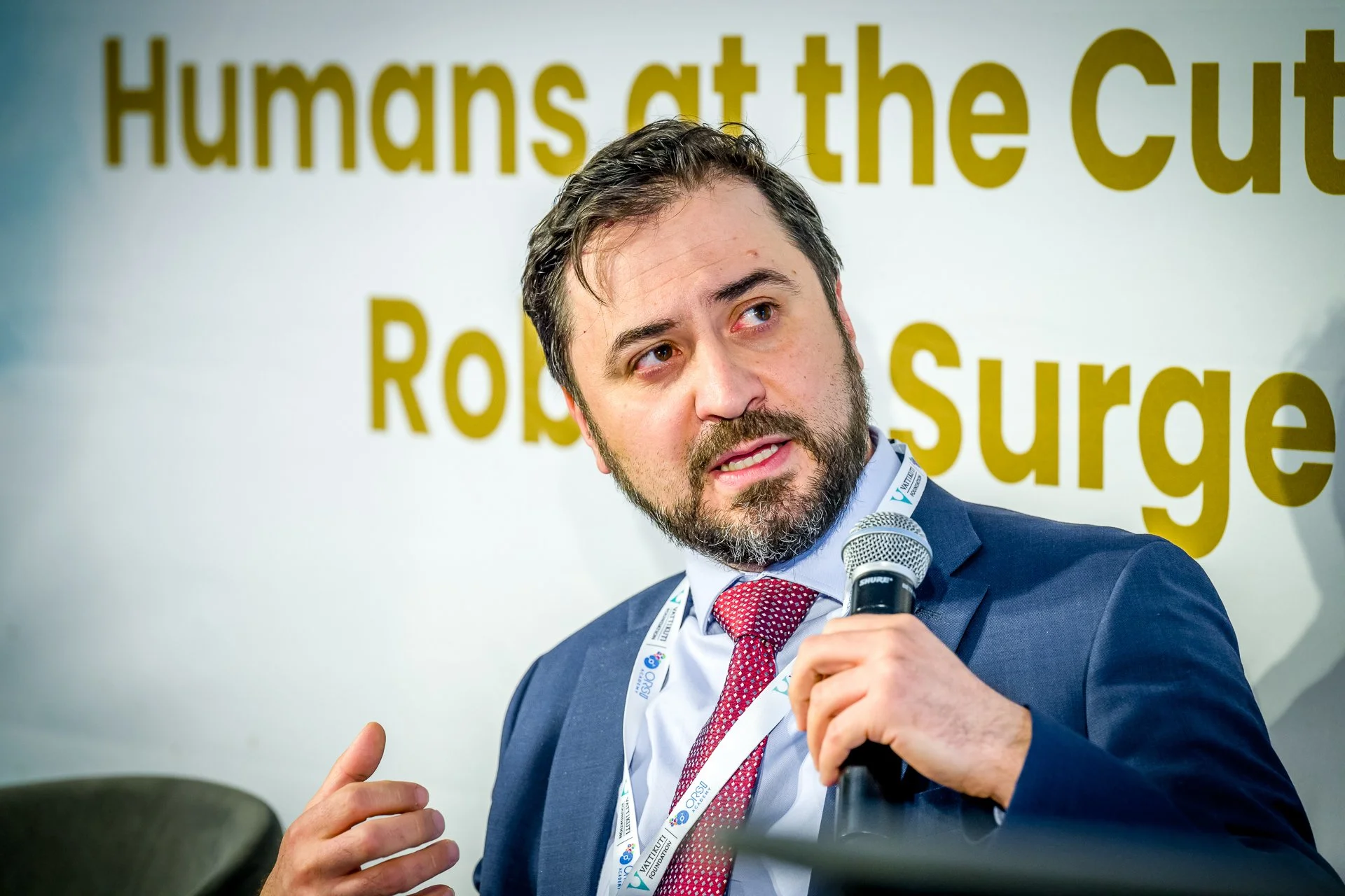 Un homme en costume tenant un micro, parlant devant un arrière-plan avec le texte "Humans at the Cutting Edge Robot Surgery".