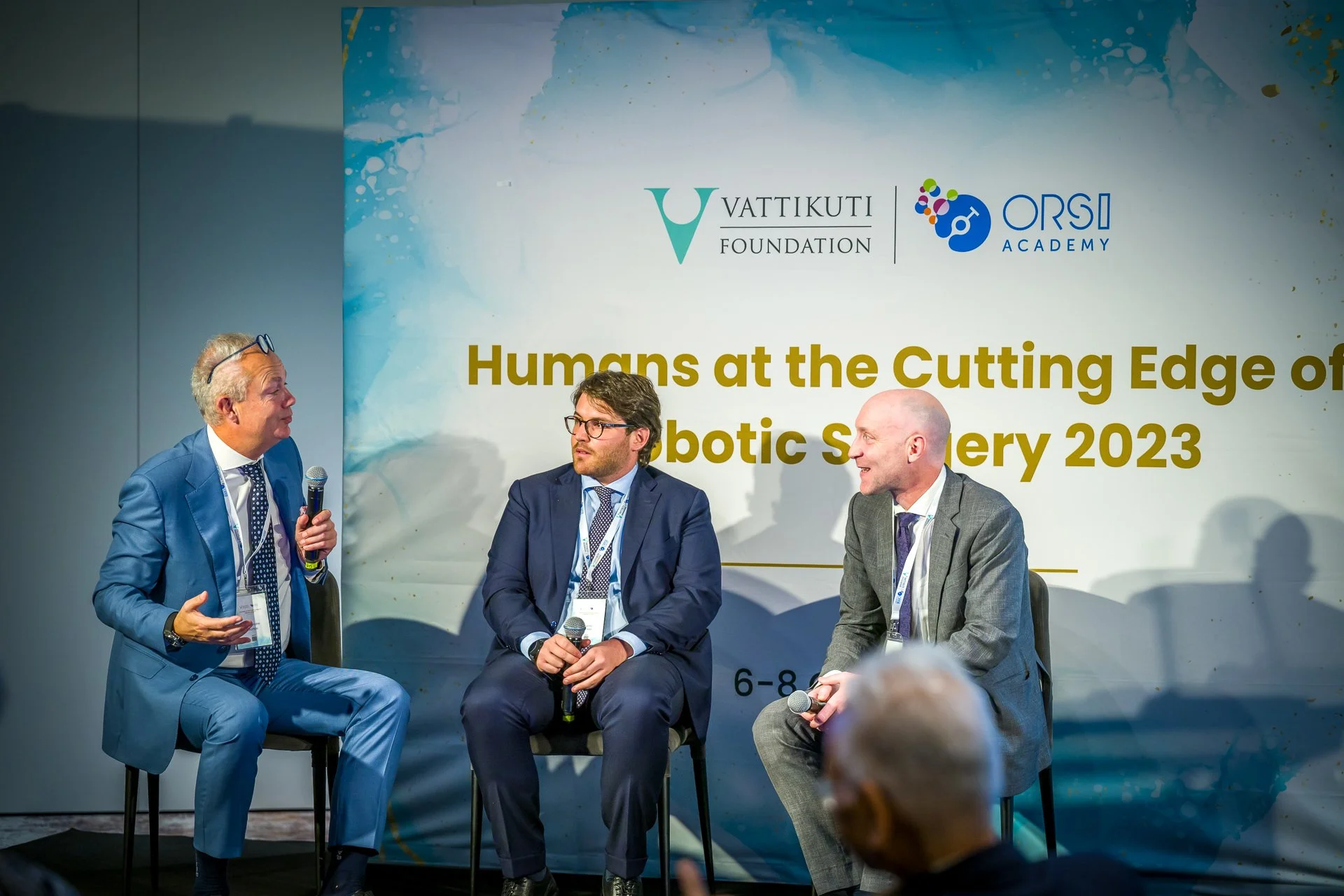 Trois hommes en costume discutent lors d'une conférence intitulée "Humans at the Cutting Edge of Robotic Surgery 2023" avec des logos de la Vattikuti Foundation et de l'ORSI Academy en arrière-plan. Les participants tiennent des microphones.