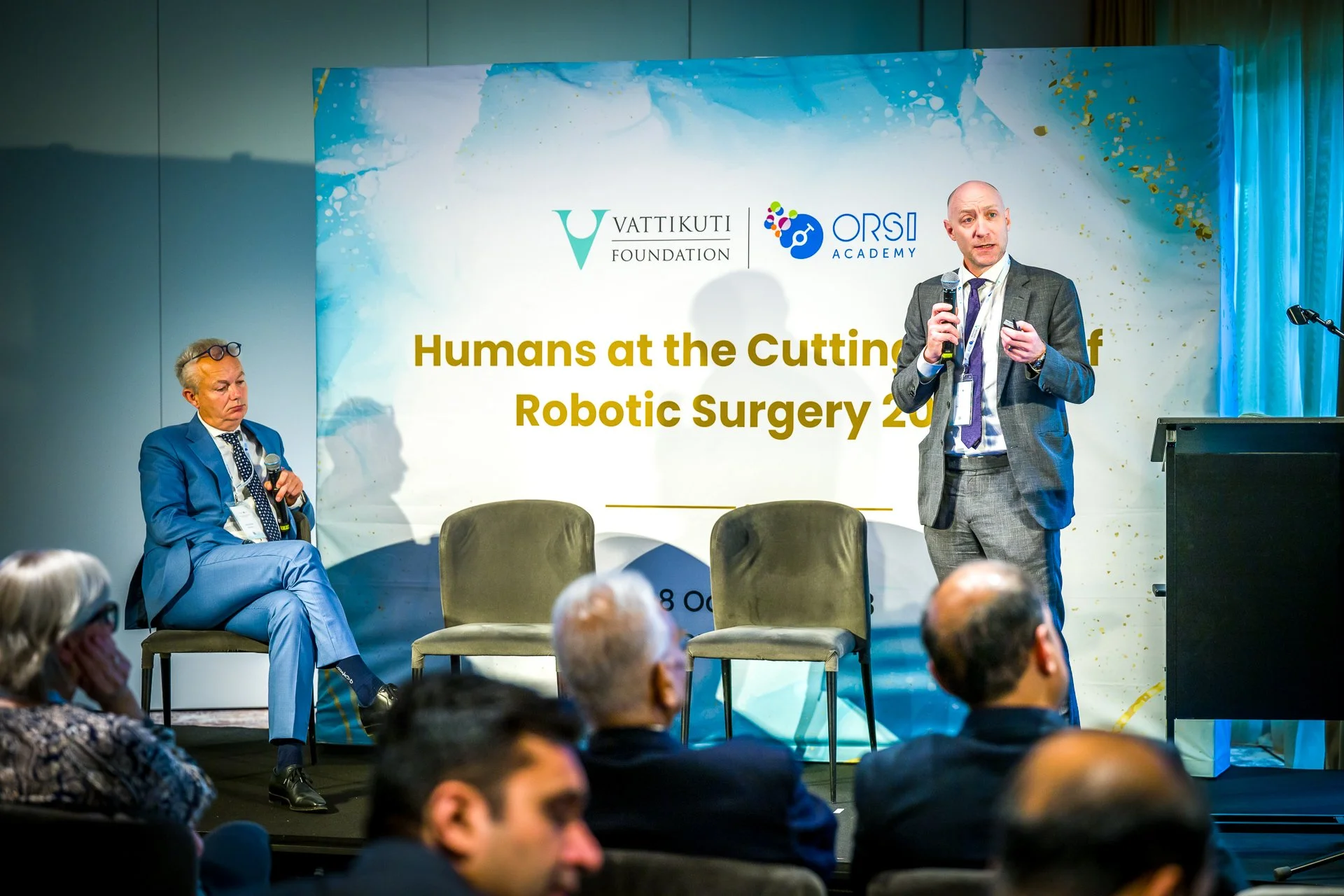Deux hommes en costume discutent lors d'une conférence sur la chirurgie robotique, avec un fond d'affiche indiquant "Humans at the Cutting Edge of Robotic Surgery 2023". Un homme est assis avec un microphone et l'autre debout parle au public.