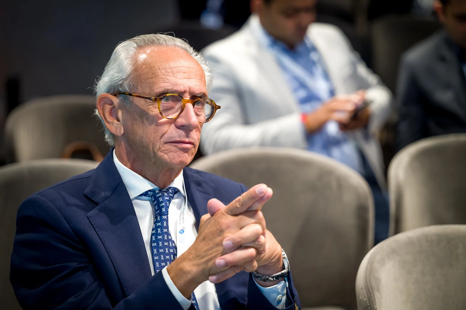 Un homme âgé portant un costume et des lunettes est assis dans une salle de conférence, les mains jointes. En arrière-plan, une autre personne est occupée sur un appareil mobile.