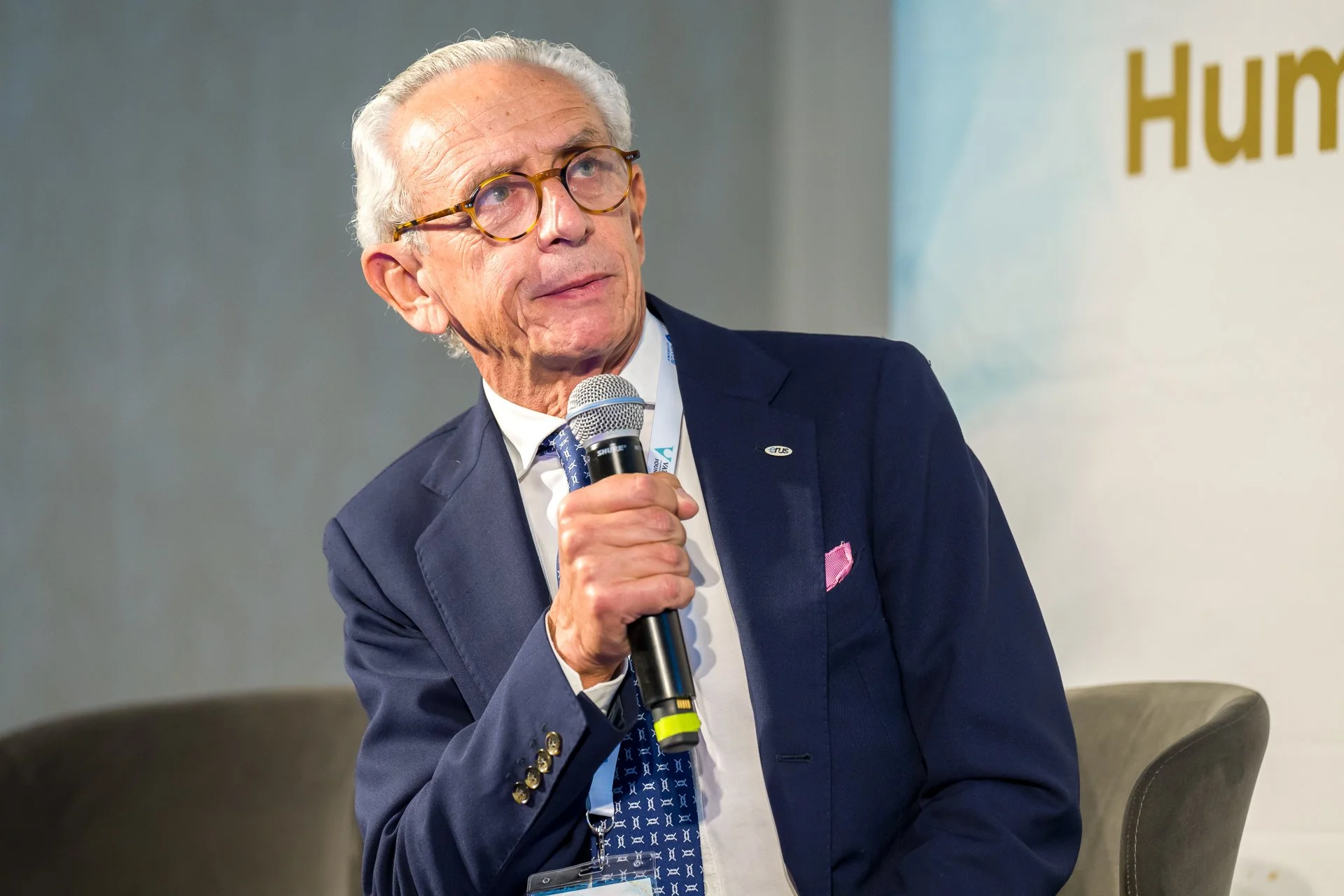 Un homme âgé en costume bleu parle dans un micro lors d'une conférence ou un événement.