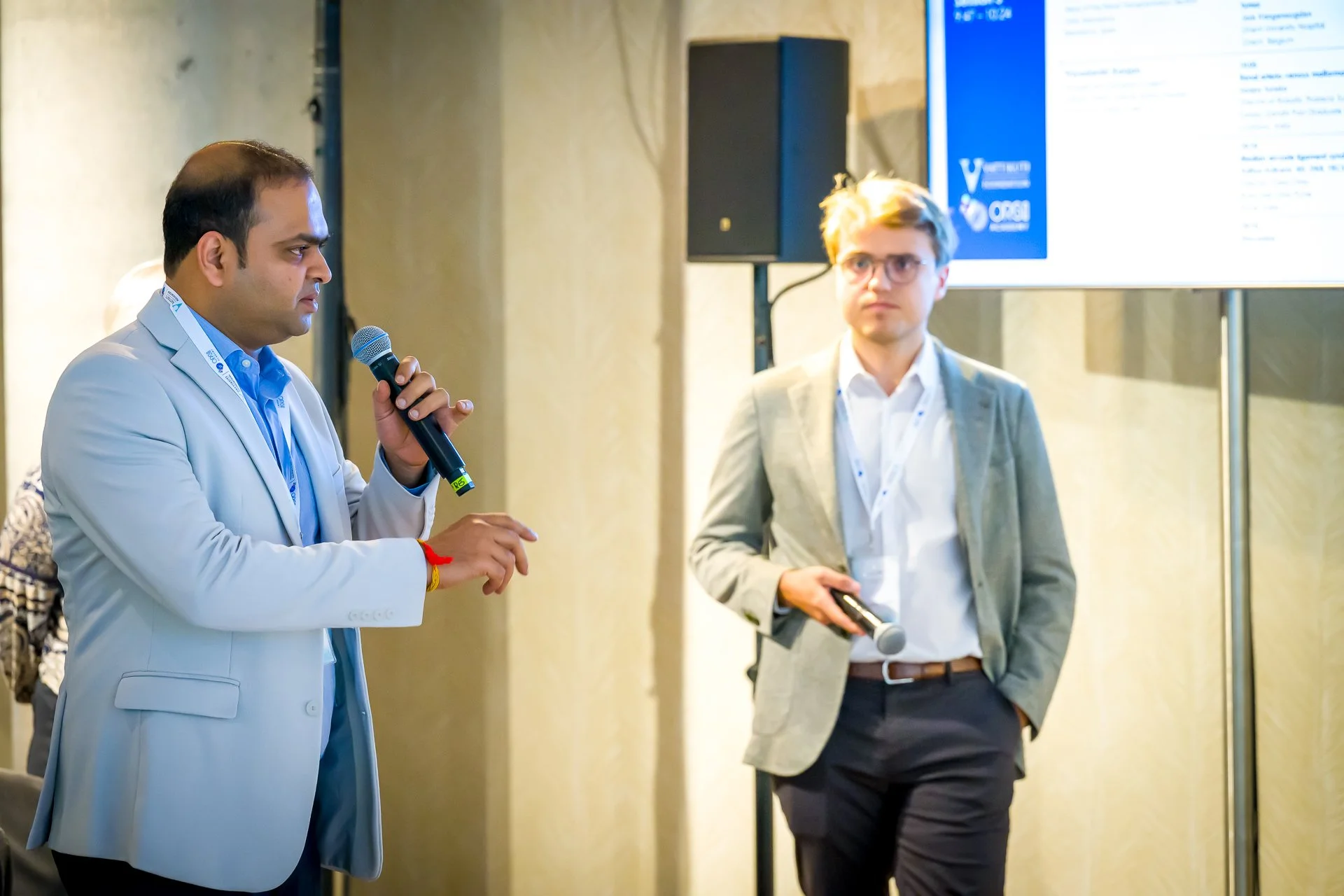 Deux hommes en costumes tenant des microphones lors d'une présentation professionnelle.