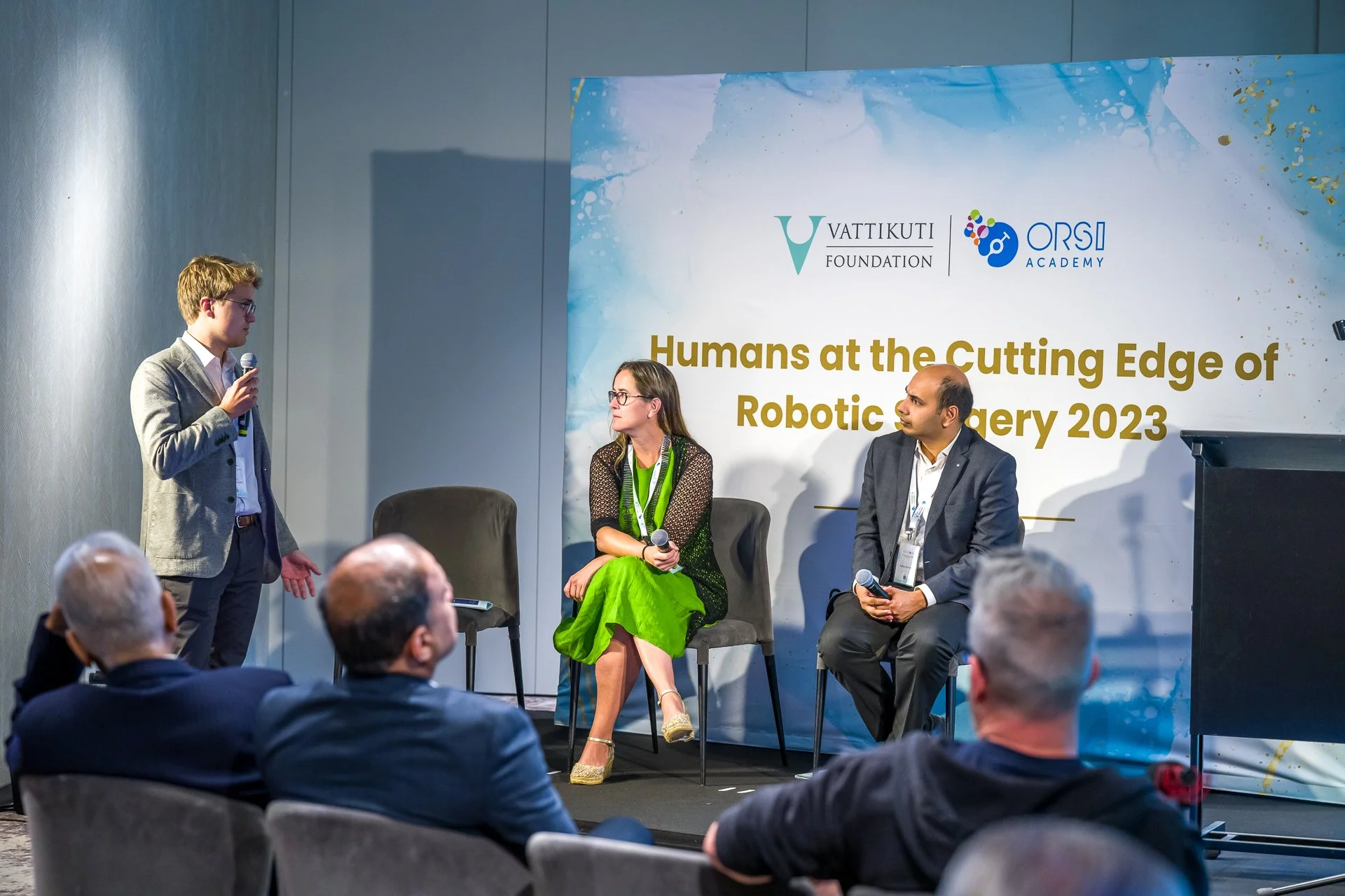 Des personnes participant à une conférence sur la chirurgie robotique, avec un homme tenant un micro et deux autres personnes assises sur scène, devant une affiche indiquant « Humans at the Cutting Edge of Robotic Surgery 2023 ».