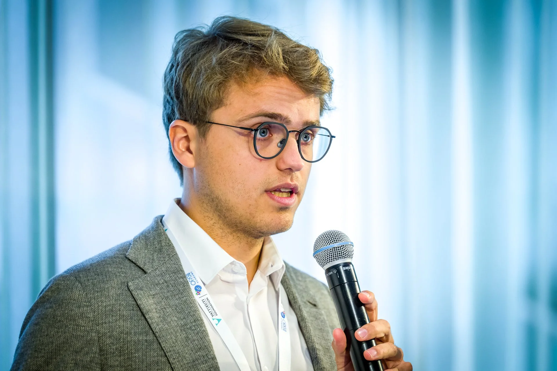 Homme avec lunettes, tenant un microphone, portant un costume gris et une chemise blanche, parlant dans un cadre de conférence.