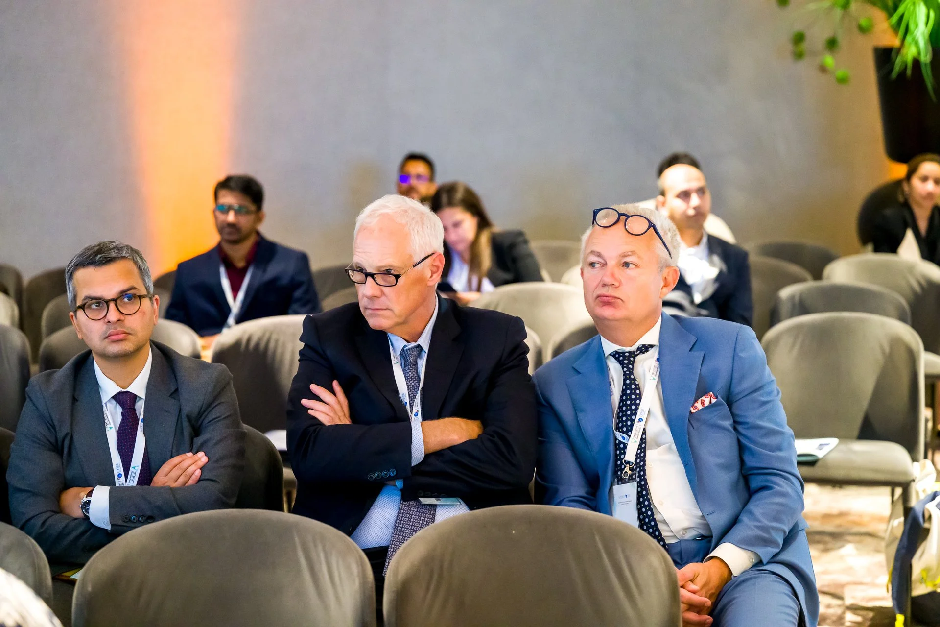 Des personnes assises dans une conférence, portant des costumes, semblent attentives et concentrées.