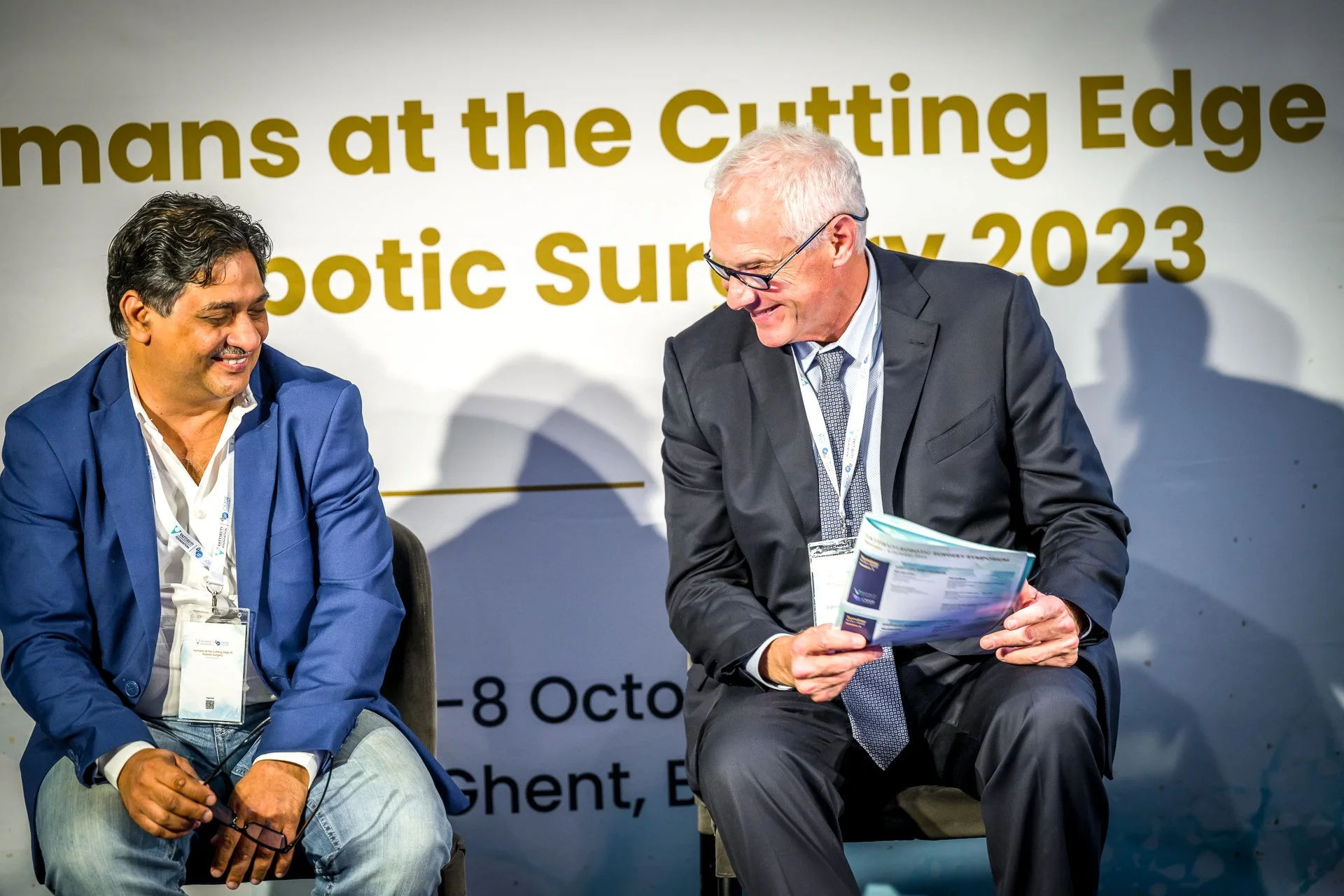 Deux hommes en costume assis sur une scène lors d'une conférence, souriant et discutant. En arrière-plan, une affiche sur la chirurgie robotique en octobre 2023 à Gand, Belgique.