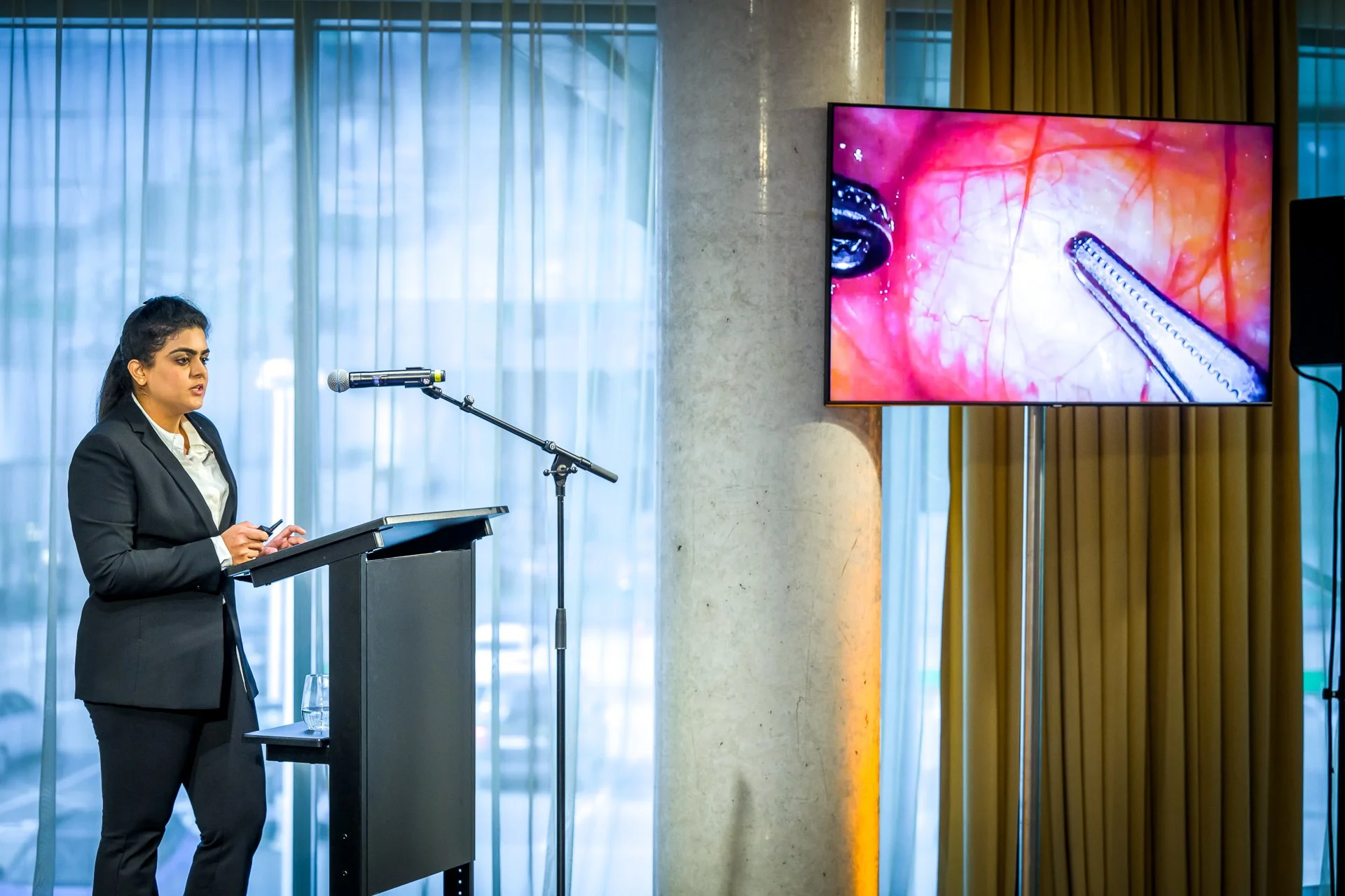 Une personne en costume noir présente à un pupitre avec un microphone. À droite, un écran affiche une image médicale, probablement une vue d'une chirurgie interne, avec des instruments chirurgicaux visibles.