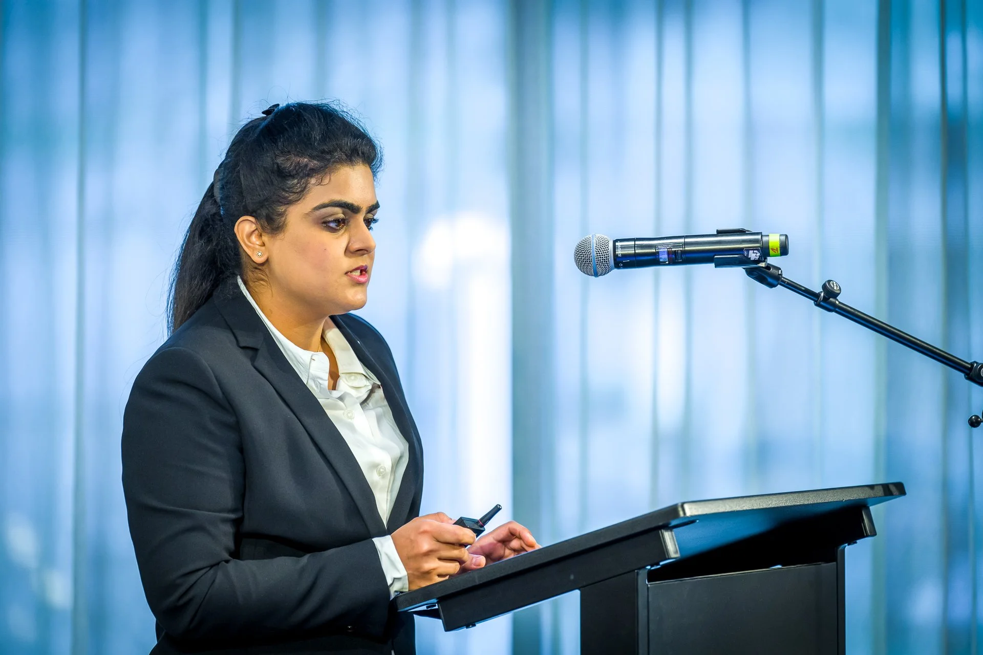 Femme en costume parle à un micro lors d'une présentation, tenant un dispositif électronique.