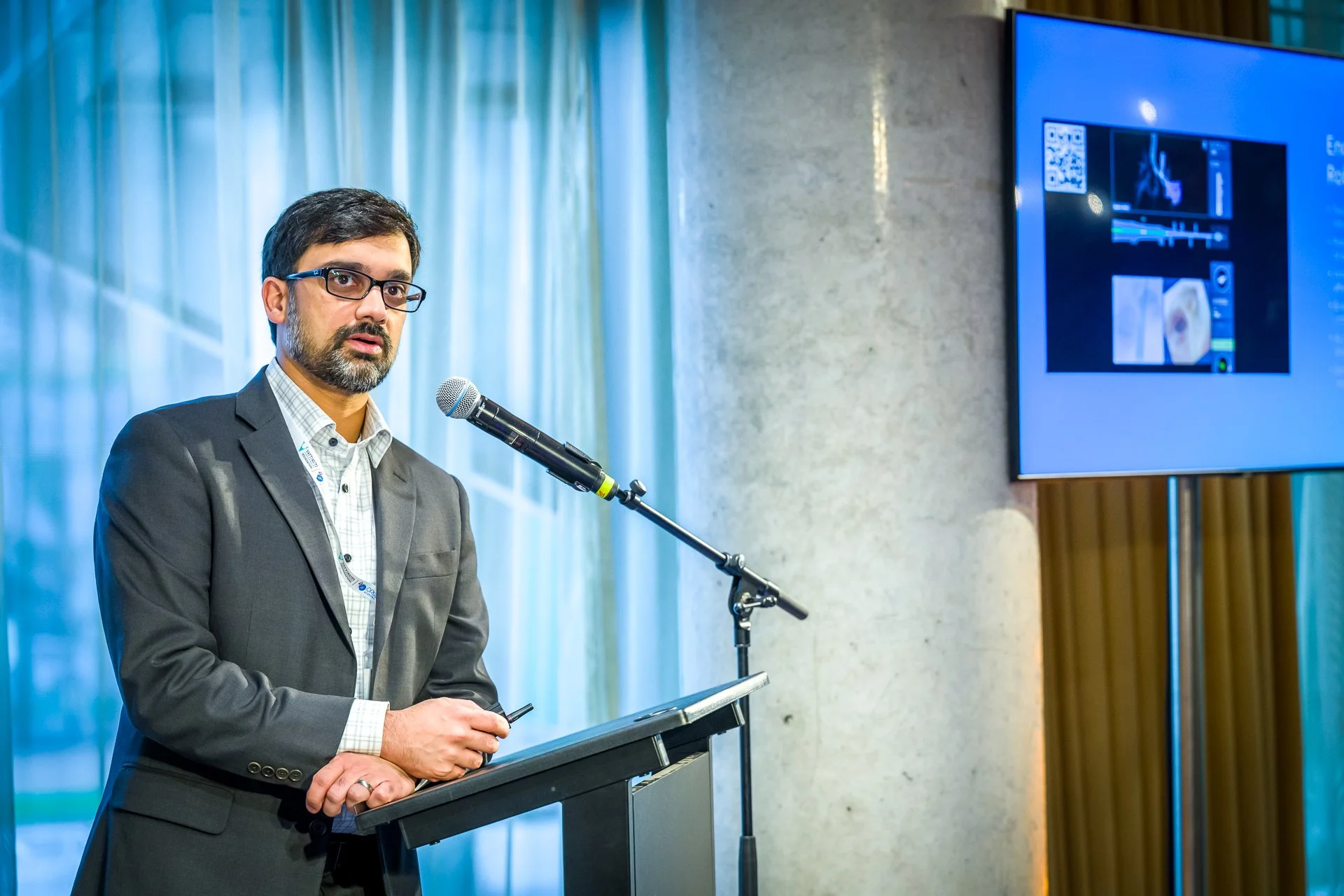 Un homme avec une barbe et des lunettes parle devant un microphone lors d'une conférence. Il porte un costume sombre et une chemise à carreaux. À l'arrière-plan, un écran présente des images et des graphiques.