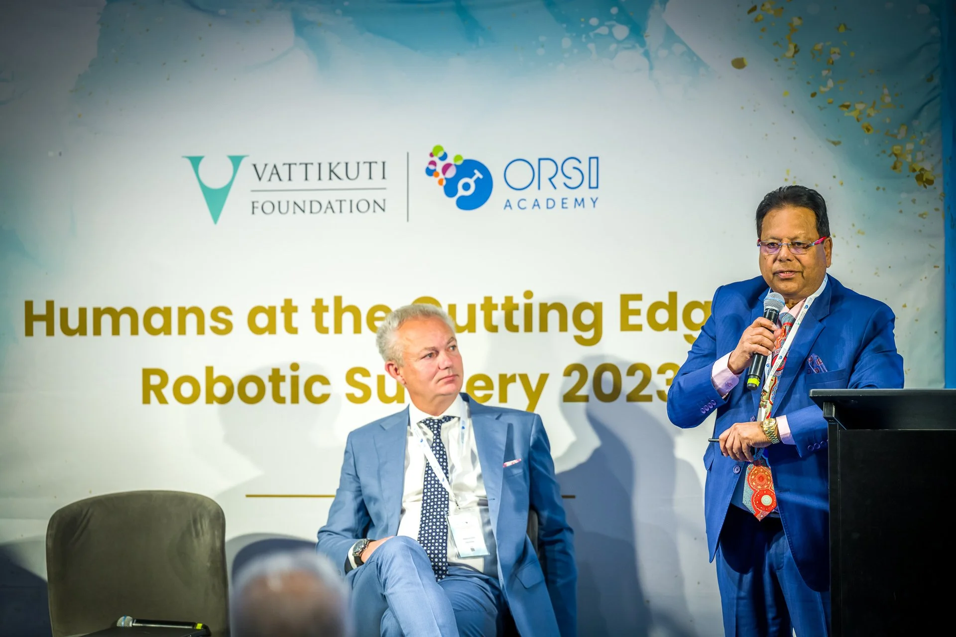 Deux hommes en costume participent à une conférence sur la chirurgie robotique, avec un écran affichant "Humans at the Cutting Edge of Robotic Surgery 2023" et les logos de la Vattikuti Foundation et de ORSI Academy.